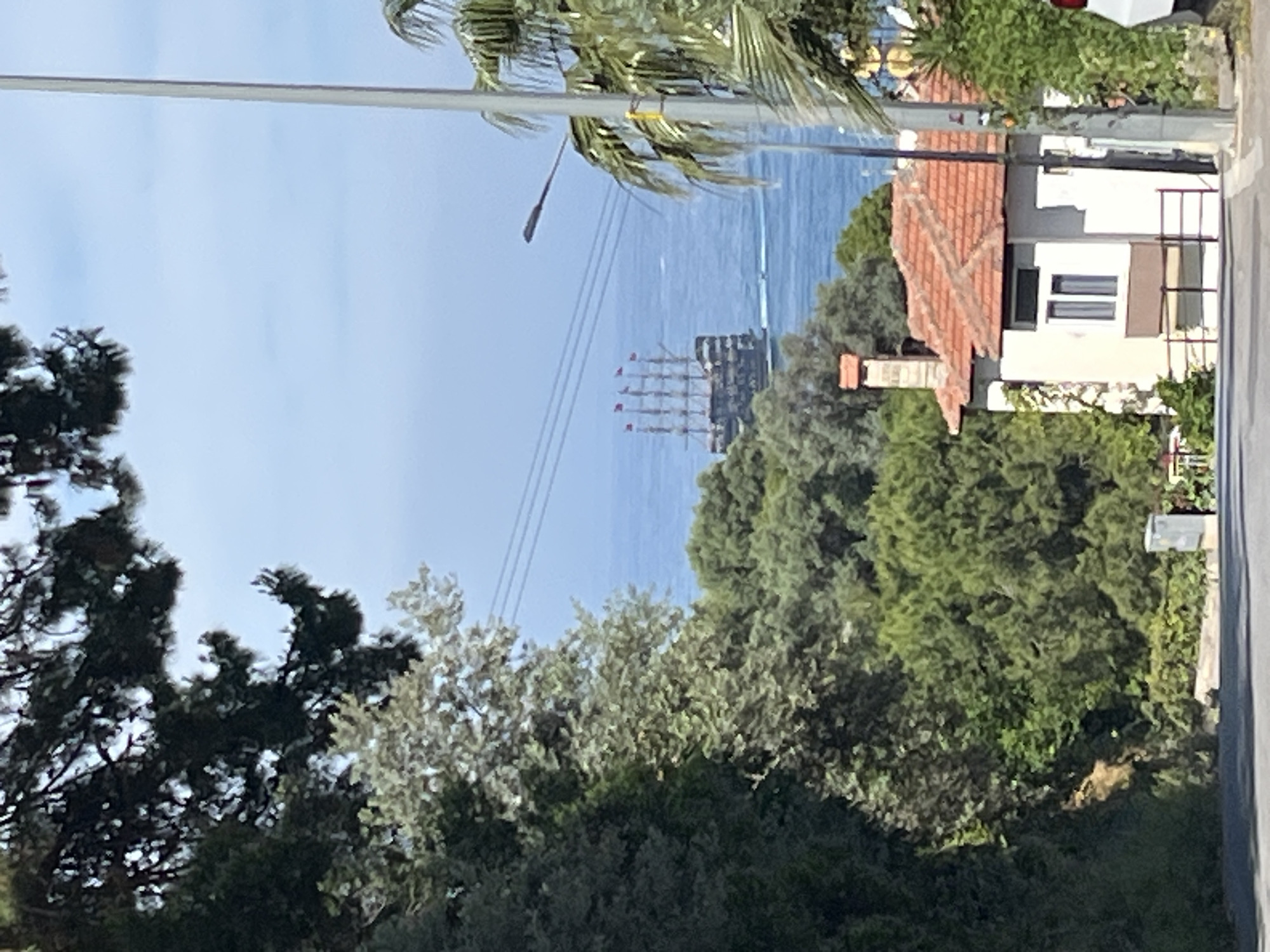 The image depicts a view towards the sea from a location in Alanya, Turkey. Visible in the midground is a large sailing ship with multiple masts, flying red flags, on the water. The foreground is dominated by lush green trees and foliage, with glimpses of buildings and infrastructure, including a house with a red tile roof and a white exterior, and telephone poles. The sky is blue with some wispy clouds. The overall scene suggests a coastal residential area with a view of maritime activity.