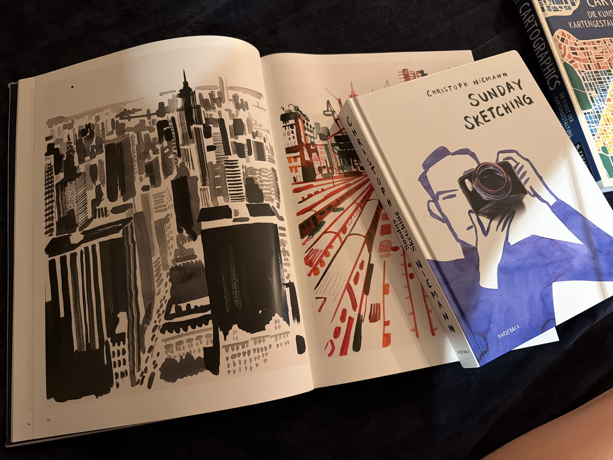 The image shows an open book with ink sketches on both pages. The left page features a black and gray abstract ink drawing of what appears to be a cityscape or architectural elements. The right page contains a vibrant ink sketch with red and green lines radiating from a central point, possibly representing a stylized explosion or burst. Resting on the open book is a hardcover book titled "SUNDAY SKETCHING" by CHRISTOPH NIEMANN, which has a cover illustration of a person holding a camera. Another book titled "CARTOGRAPHICS" with the subtitle "DIE KUNST DER KARTENGESTALTUNG" is visible to the right, displaying a colorful map-like illustration. The books and the open sketchbook are placed on a dark, soft surface, likely fabric, casting shadows. There is no indication of the city of Mühlbach - Rio di Pusteria, Italy within the image.