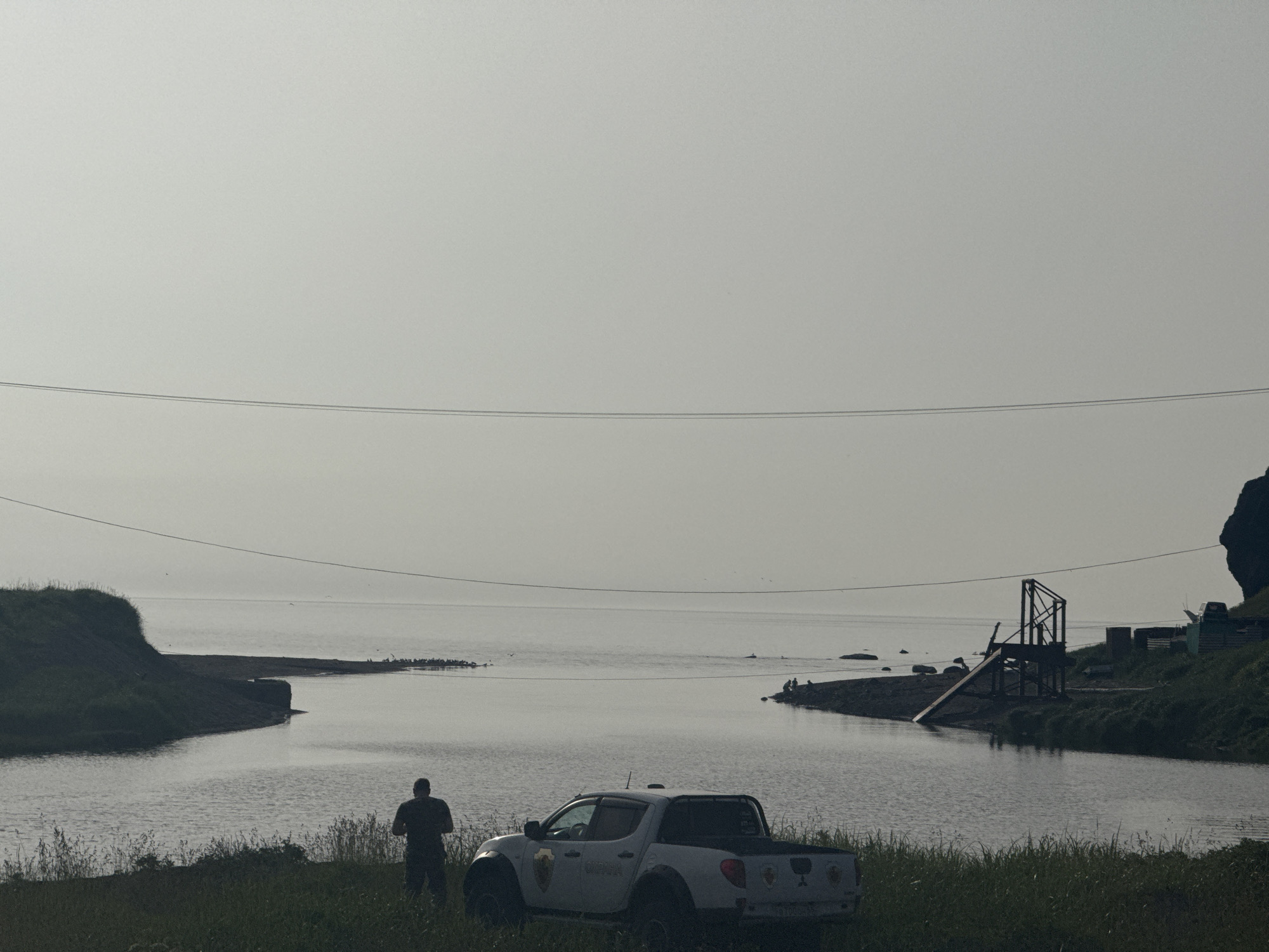 The image shows a gray, overcast day at a coastal location, possibly in the city of Kuril'sk, Russia. In the foreground, a white pickup truck is parked on a grassy embankment. A person, silhouetted against the light, stands near the truck looking out at the water.
The scene opens up to a calm body of water, likely a bay or inlet, with a gently curving shoreline. On the opposite side of the water, there is a man-made structure resembling a ramp or a small dock, with some additional structures visible further inland. Several small figures are seen sitting or standing near this structure, possibly engaged in fishing or other activities. The horizon line is indistinct due to the overcast conditions, with the sky and water blending together. Two power lines stretch horizontally across the upper portion of the frame, adding a man-made element to the natural landscape. The overall impression is one of a quiet, somewhat remote coastal setting.