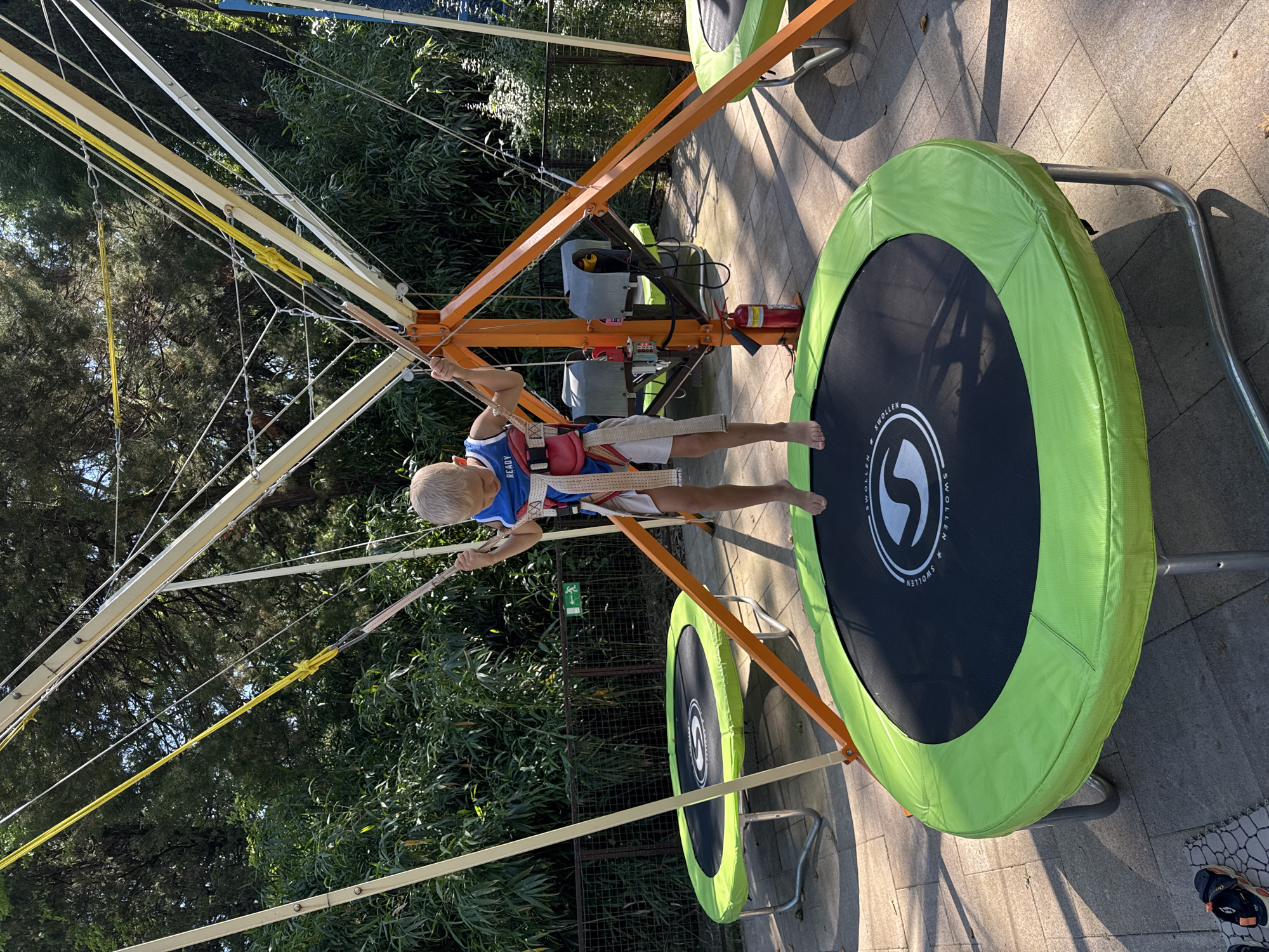 A young boy is tethered by bungee cords and a harness to a metal and orange frame, positioned over a trampoline with a green rim. He is barefoot and dressed in a blue and red tank top with white shorts. He appears to be in mid-air, possibly jumping. Two other trampolines with green rims are visible in the background, one to the left and one to the right, partially obscured by the frame and other structures. The scene is outdoors, set against a backdrop of lush green trees and foliage. A red fire extinguisher is mounted on the orange frame. The ground is paved with interlocking stone tiles, and shadows are cast on the ground, suggesting it is a sunny day. A wire fence runs horizontally behind the trampolines, with a small green exit sign with an arrow on it visible on the fence. The overall setting suggests an amusement park or recreational area in Sochi, Russia.