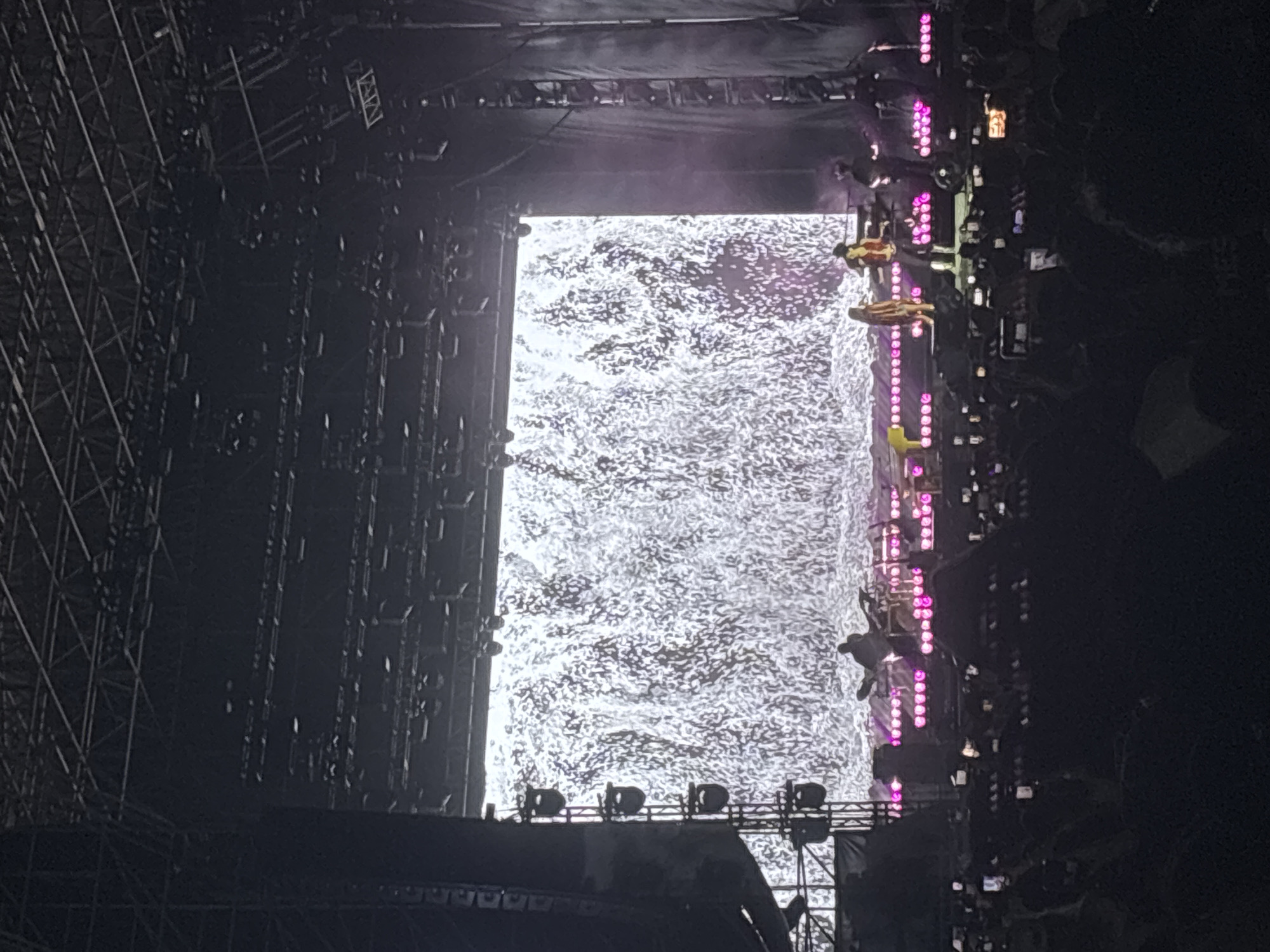 The image depicts a concert stage in Tampico, Mexico, at night. A large screen in the center displays a swirling, light-colored pattern. To the right of the screen, people are suspended from rigging, illuminated by vertical rows of pink lights. Stage scaffolding is visible on the left side of the screen. At the bottom of the screen, stage lights are positioned on a platform. Audience members are present on the right.