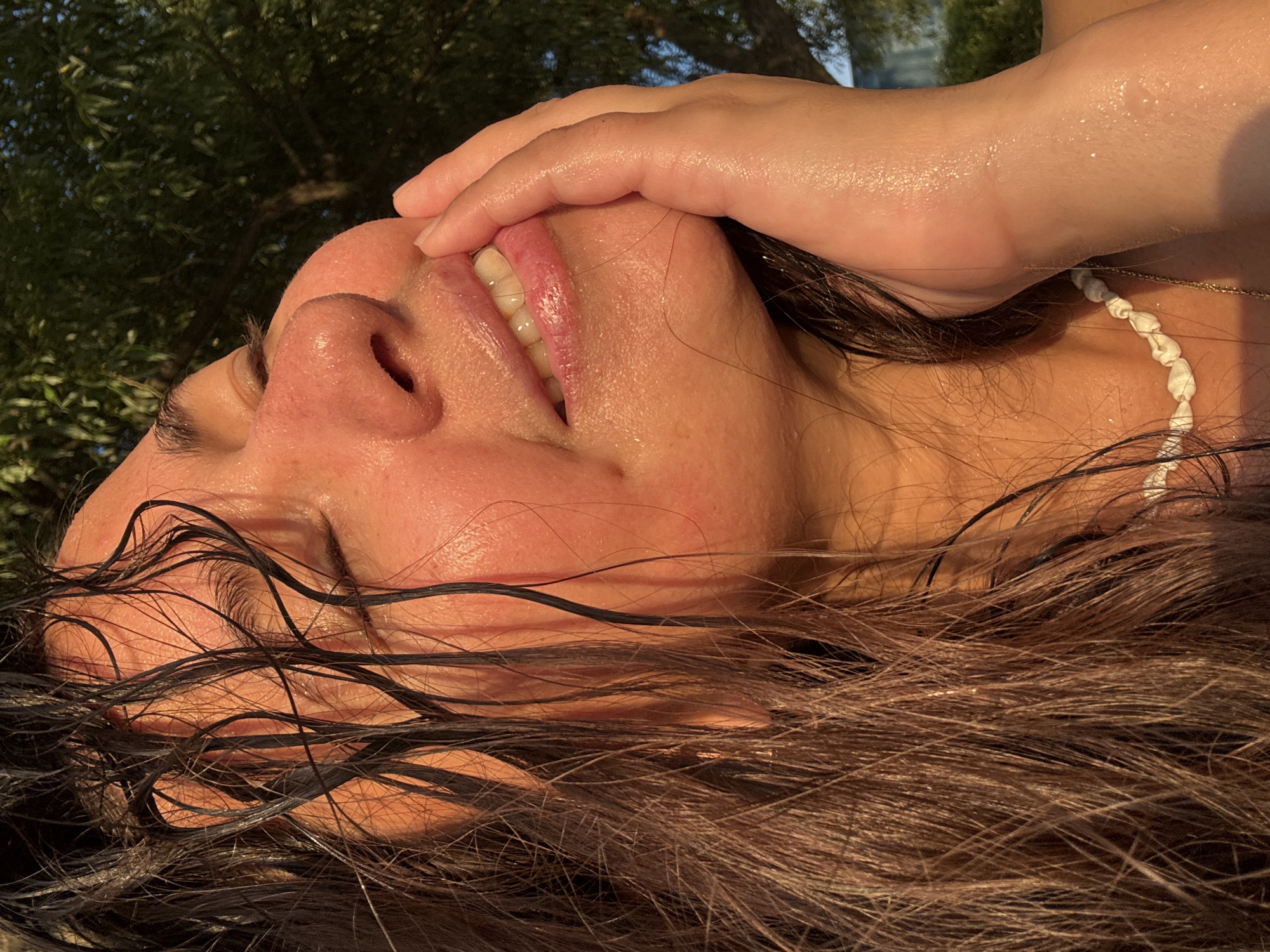 A close-up, waist-up, outdoor shot shows a woman with her eyes closed, her face turned to the right, and her left hand touching her cheek. Her wet, dark brown hair is falling around her face and shoulders. The sun illuminates her face from the side, highlighting her features and creating a warm glow. She is wearing a white shell necklace. The background is blurred, but appears to be a green, leafy tree, suggesting a natural outdoor setting. The image does not provide any information that would allow for its location to be identified as Kamysh, Russia.