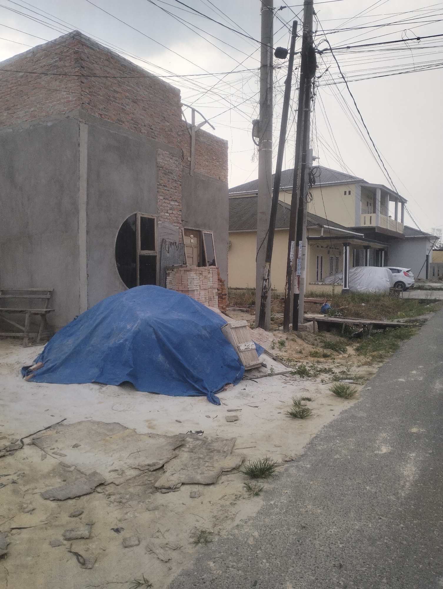 The image depicts an unfinished brick and concrete building under construction in Tambusai Batang Dui, Indonesia. In the foreground, a large pile of material is covered with a blue tarp. A wooden bench is visible to the left of the tarp. The building features exposed brickwork on the upper section and concrete on the lower section, with a circular window opening. Adjacent to the building, there is a stack of bricks and some window frames. Further in the background, a light yellow two-story house with balconies and a gray house are situated. A white car is parked in front of the yellow house, covered with a light-colored tarp. Utility poles with numerous electrical wires are prominent in the scene. The ground is a mixture of dirt, sand, and some patchy grass. The overall environment appears to be a residential area undergoing development.