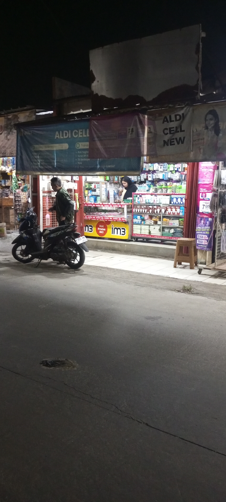 This is a vertical, medium-shot, nighttime photograph taken on a street in front of a store in Setu, Indonesia. The store is illuminated and appears to be a small convenience store or cell phone shop. Above the storefront, a blue banner reads "ALDI CELL NEW". Inside the store, shelves are stocked with various products. A woman wearing a black headscarf stands behind the counter, facing away from the camera, and a man in a dark jacket walks towards the store entrance. In the foreground, to the left, is a black motorcycle parked on the side of the road. The road surface in the foreground is asphalt and shows signs of wear, including a pothole and cracks. The environment appears to be urban, with the store being the primary source of light in the scene.