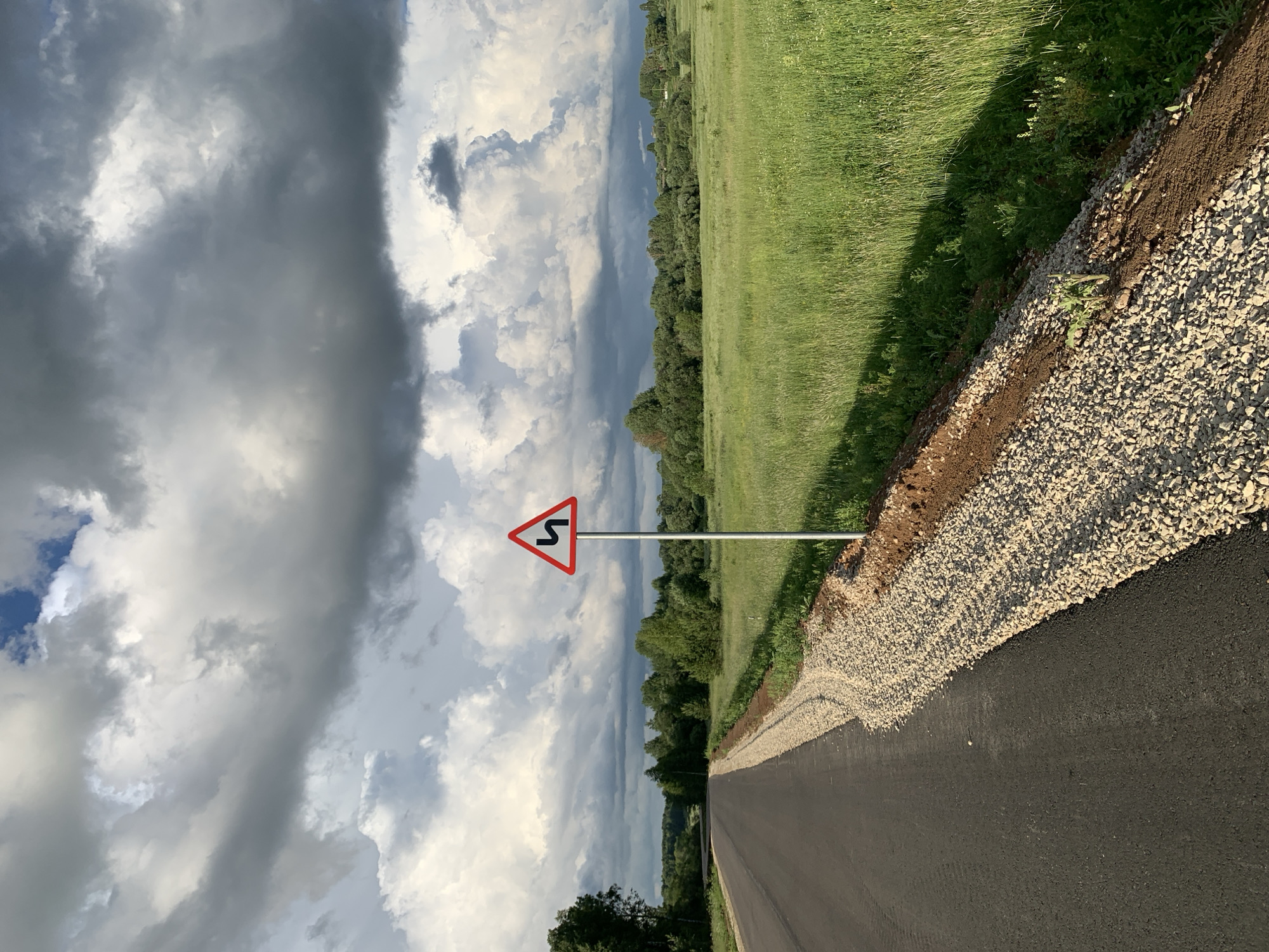 This is a photograph of a rural road in the city of Svyatitsa, Russia. The road is paved and curves to the left, with a gravel shoulder on the right. A traffic sign warning of sharp curves ahead is visible on the right side of the road. The surrounding landscape is a green field with trees in the distance. The sky is filled with dramatic, dark clouds.