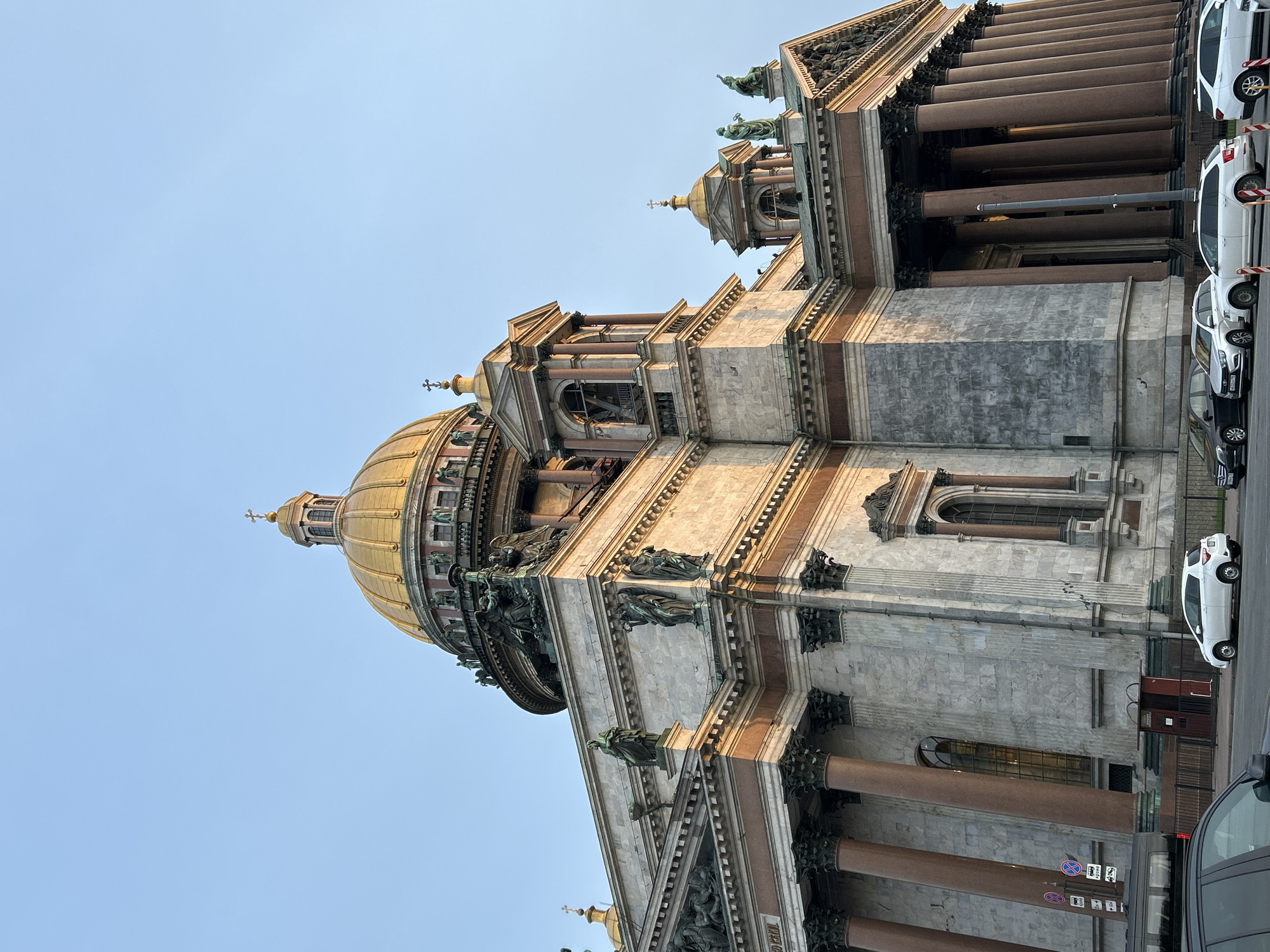 The image depicts a low-angle, side view of St. Isaac's Cathedral in Saint Petersburg, Russia. The cathedral's exterior is composed of gray stone and bronze, with a large golden dome. Classical columns line the building's facade. Statues and ornamentation are visible on the structure. A row of parked cars is seen on the side of the street next to the building. The sky is blue.