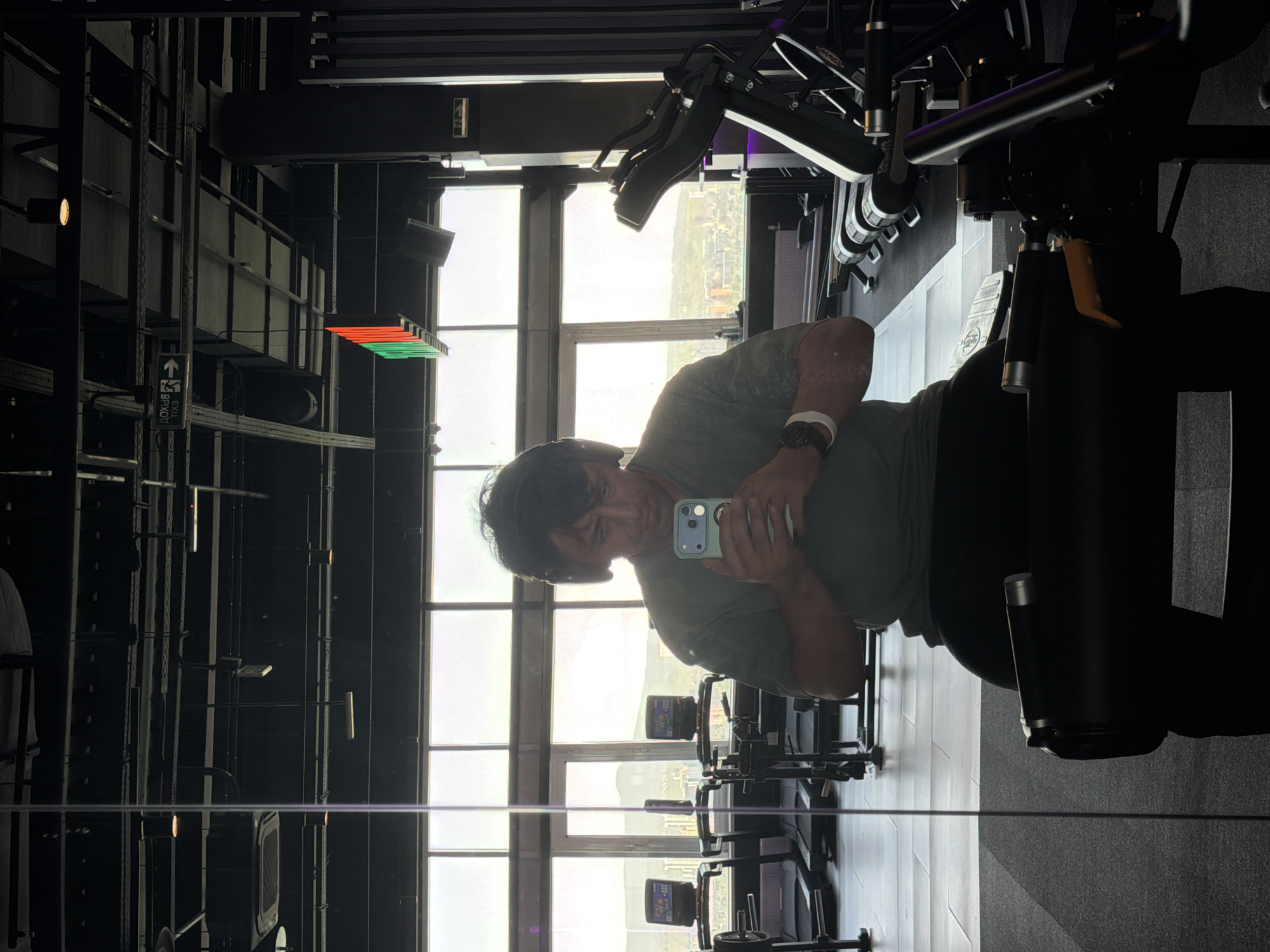 A person is captured taking a mirror selfie in a modern gym or fitness center, oriented ninety degrees clockwise in the image. The man, wearing a green t-shirt and a watch on his left wrist, holds a smartphone with a light blue-green case to take the picture.
The setting is a spacious, well-lit gym featuring large floor-to-ceiling windows that reveal a hazy cityscape or landscape in the distance, indicating a high-level view. The gym's interior has a contemporary, industrial design with exposed dark metal beams and structures. The floor is highly reflective, showing a distorted reflection of the gym equipment and the man's lower body.
Various pieces of dark-colored gym equipment are visible, including what appears to be a leg press or similar resistance machine prominently in the foreground and blurred equipment in the background and reflection. One machine has the brand "STARTRAC" visible. Artificial lighting fixtures are present, with one bright spot on the left wall and a horizontal array of red and green lights further back.
A green sign on a dark wall features an arrow pointing to the right (when the image is correctly oriented) and Cyrillic text that reads "ВХОД" (Entrance), with another partial word that might be "ПРОХОД" (Passage). The bright light from the windows suggests it is daytime. The overall impression is a modern fitness facility with an expansive view.