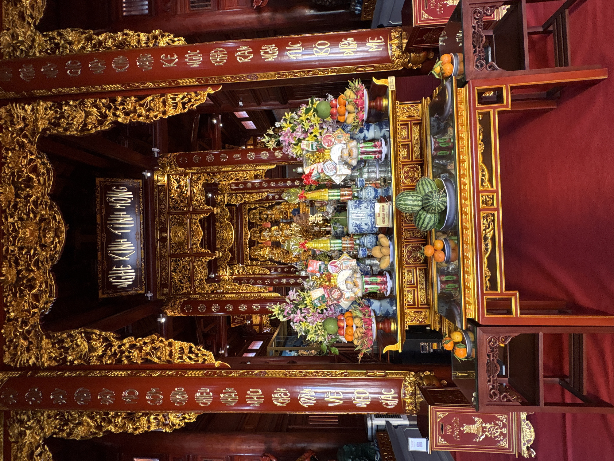 The image captures an ornately decorated space, likely a temple or religious site in Nam Nha Trang Ward, Vietnam. The scene is dominated by wooden structures, richly adorned with intricate gold detailing. The structures appear to be arranged as a display or altar, laden with offerings.
These offerings include an assortment of fruits such as oranges and watermelons, flowers, and other objects carefully arranged on multiple tiers. The use of red and gold colors throughout the scene contributes to the overall sense of opulence and reverence.
Inscriptions in a foreign script are visible on the wooden surfaces, further emphasizing the cultural and religious significance of the location. The scene is viewed from a low angle, further exaggerating the height and grandeur of the structures.