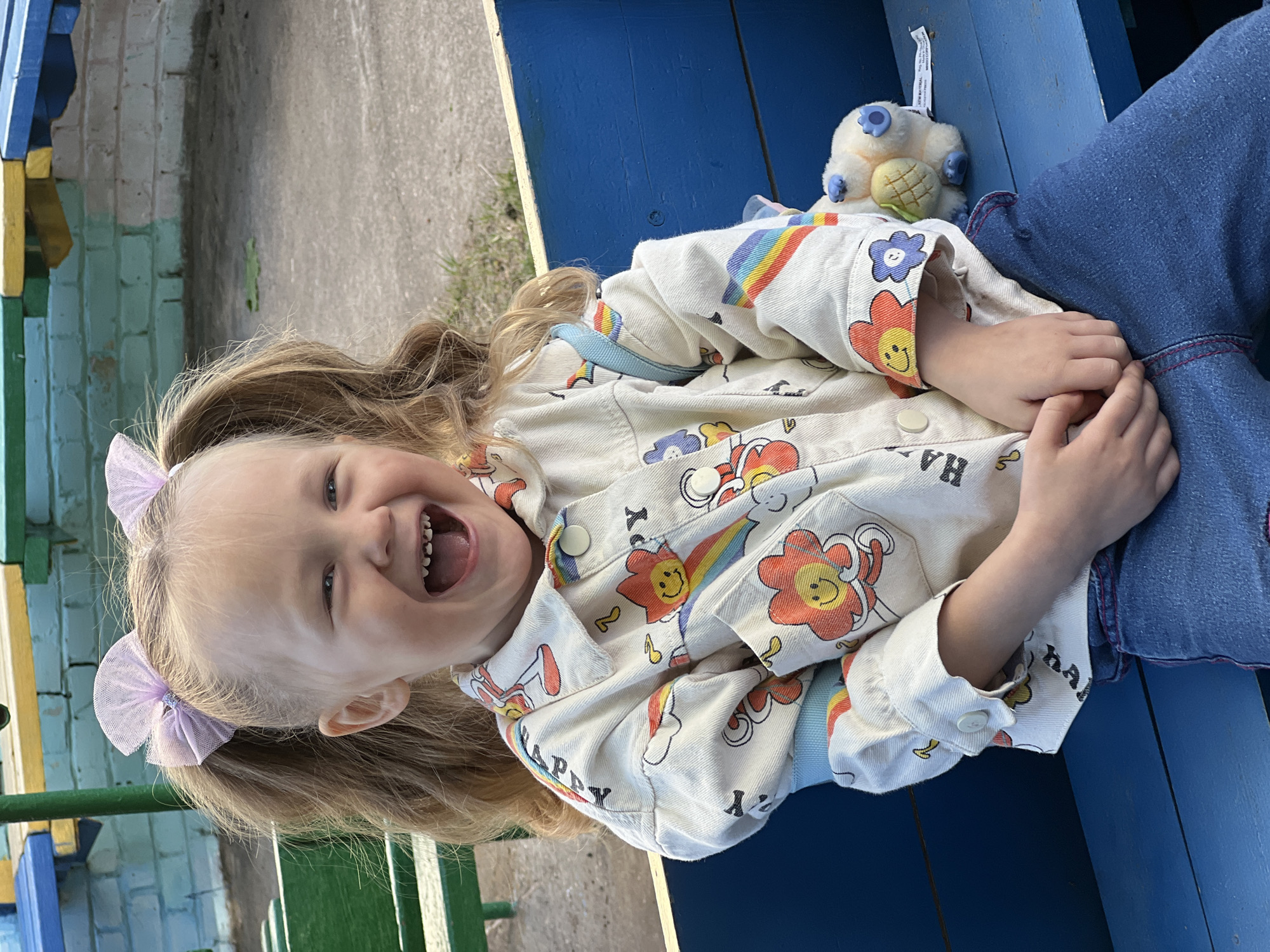 A young child with blonde hair pulled into pigtails adorned with lilac bows is laughing while sitting on a blue wooden structure. The child wears a white jacket patterned with colorful smiling suns, clouds, and rainbows, over blue jeans. A stuffed animal with a blue flower is next to the child. The location appears to be outdoors, near a painted structure with green, yellow, and blue elements. The scene may be in the city of Chaikovsky, Russia.