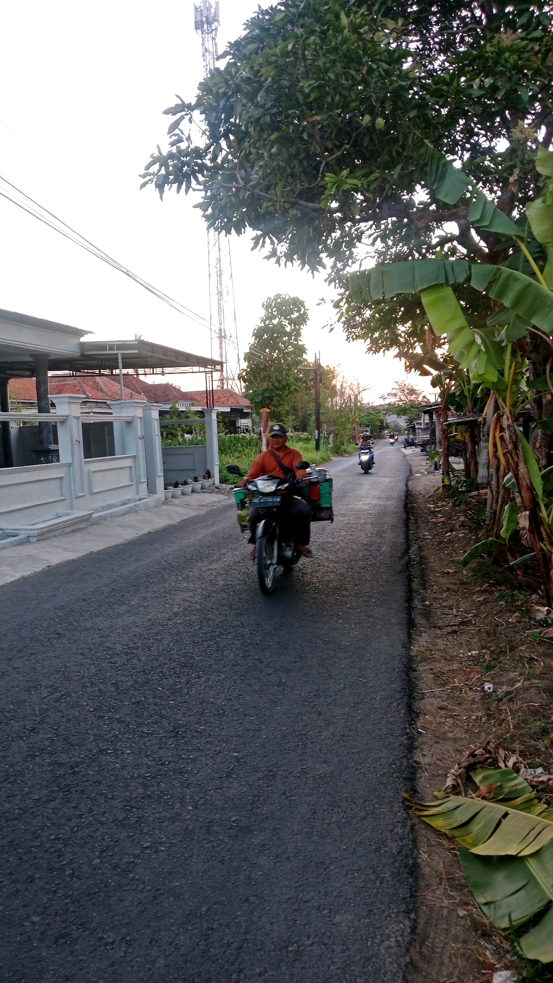 A man wearing an orange shirt and a dark cap rides a motorcycle on a paved road in Dempo Barat, Indonesia. The motorcycle has a green cargo box attached to the back. He is moving away from the camera, towards the right side of the frame. The road is bordered on the left by a white fence and the roofs of houses, and on the right by a dirt shoulder with banana trees and other vegetation. In the background, a cell tower is visible against a bright sky. Another motorcycle is visible further down the road, also moving away from the camera. The overall scene appears to be a rural or suburban residential area during daylight hours, possibly late afternoon given the lighting.