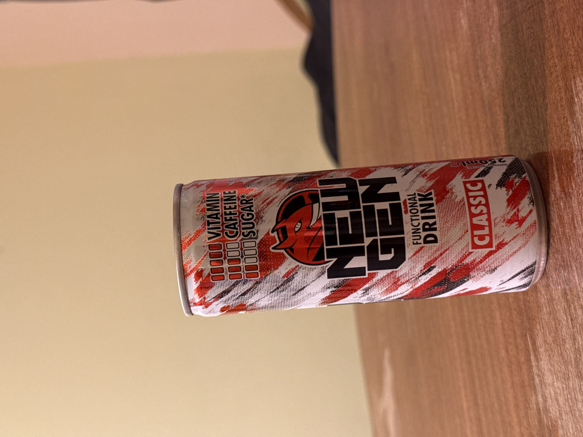 A slightly overexposed, medium-shot photograph shows a can of New Gen Classic functional drink on a wood surface, likely a table. The can features a white background with red and black streaks, alongside the New Gen logo, which includes a stylized devil's head. Text on the can lists "VITAMIN," "CAFFEINE," and "SUGAR," followed by "FUNCTIONAL DRINK," and "CLASSIC." The can is printed with "250ml" and "Нетно количество." The setting appears to be indoors, possibly a residence or office, given the beige wall in the background and a dark, ambiguous shape that may be clothing. There is no visible indication of Stara Zagora, Bulgaria.