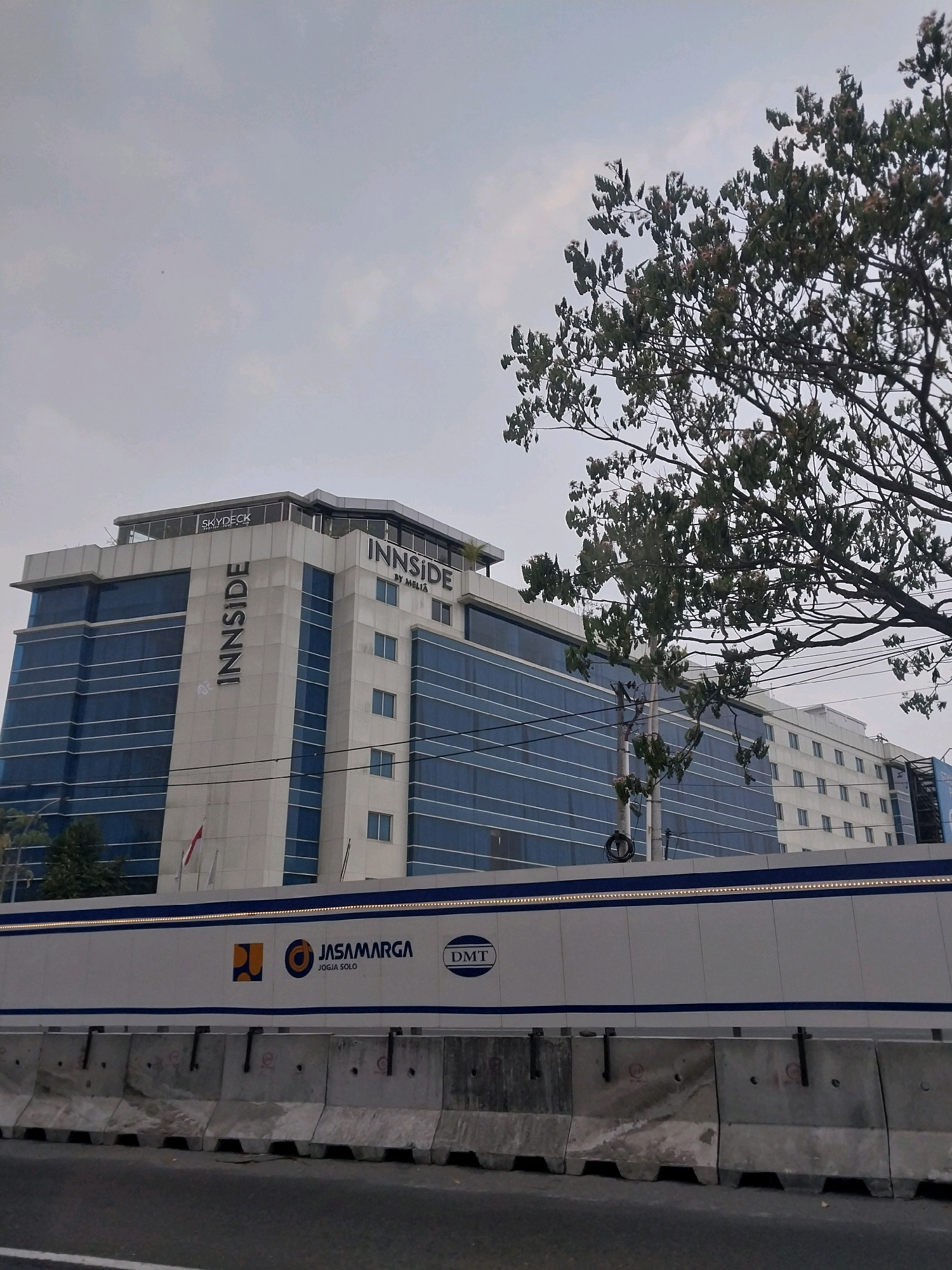 The image shows a modern hotel building with a blue glass facade and white accents. The hotel's name, "INNSIDE BY MELIA," is prominently displayed. On the roof of the hotel, the word "SKYDECK" is visible. In front of the hotel, there is a white barrier with blue accents and logos of "JASAMARGA JOGJA SOLO" and "DMT." The barrier is made of concrete blocks. A tree with green leaves is partially visible in the foreground, obscuring part of the hotel. The sky is overcast. The scene appears to be in an urban or suburban environment.