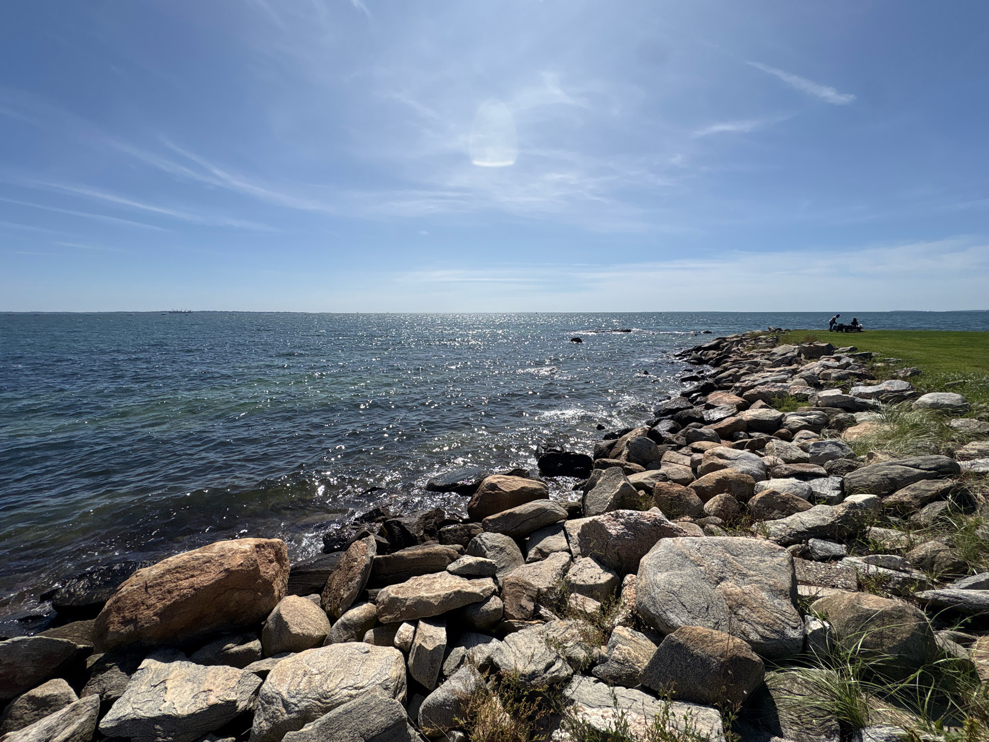 The image depicts a rocky shoreline with a body of water on the left and a grassy area on the right. The water, likely the ocean, is a deep blue with sunlight glinting off its surface. The shoreline is covered with numerous rocks of varying sizes and colors, from light gray to reddish-brown. Some dry grass and weeds are growing between the rocks. In the distance, on the grassy area, two individuals are seated on a bench, appearing to be enjoying the view. The sky above is clear blue with a few wispy white clouds. The scene appears to be a public park or waterfront area in Waterford, United States of America, offering a serene natural landscape.
