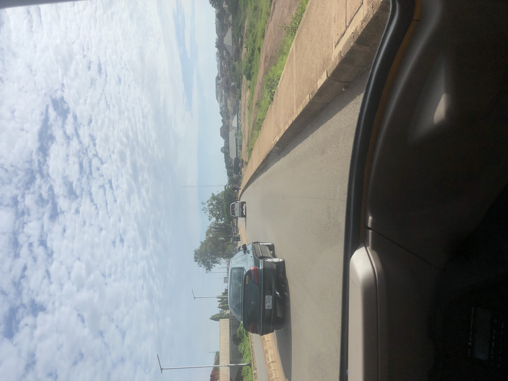 A car is driving down a road in Barakin Lerawi, Nigeria. The car is dark green and has a license plate that reads "KWL 465PN". The car is in the left lane of a two-lane road. There are other cars in front of it, as well as trees and buildings on the side of the road. The sky is cloudy and blue. The sun is shining.