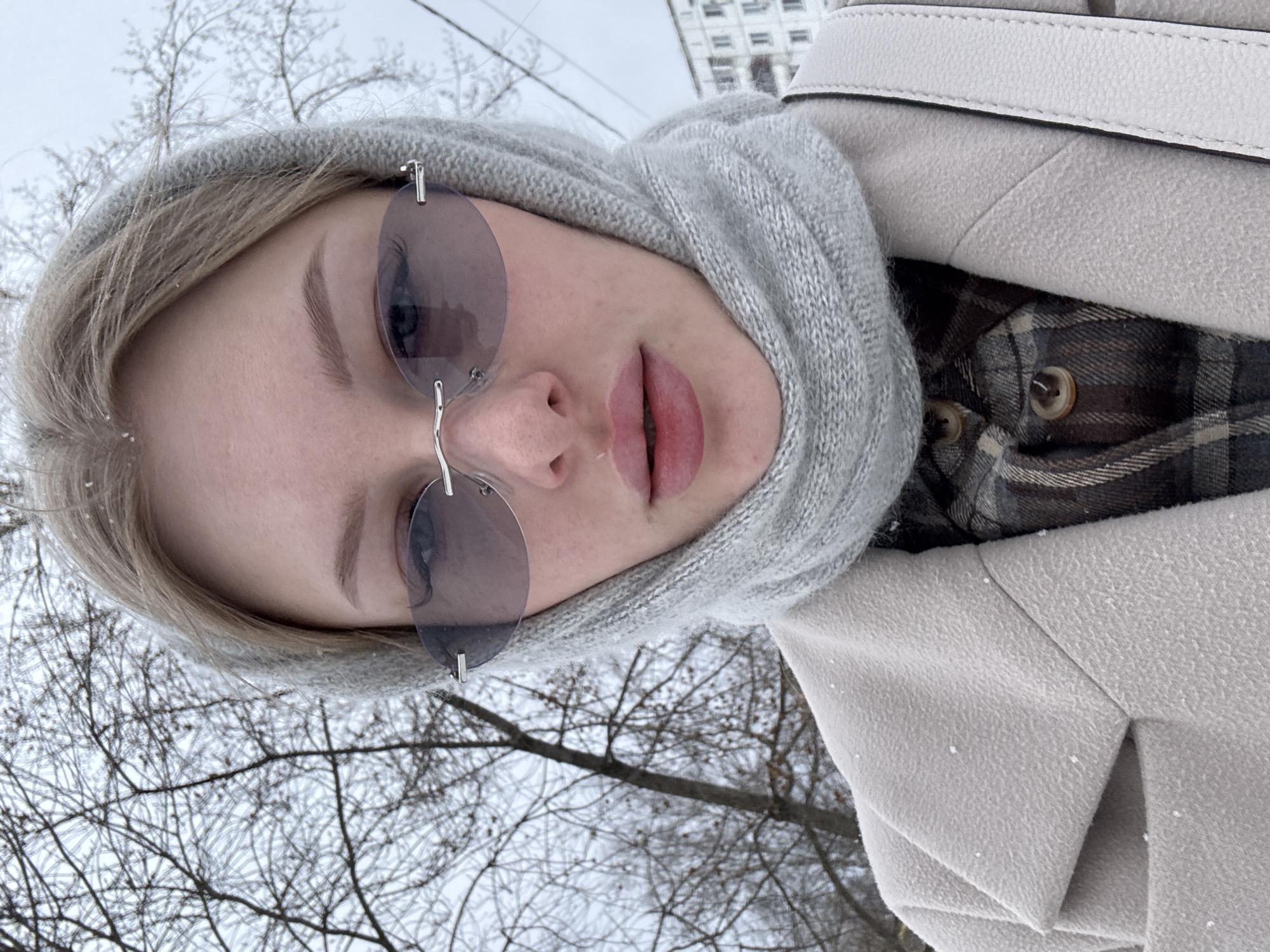 A close-up selfie captures a person outdoors in a winter setting in Perm, Russia. The individual, likely a young woman, has blonde hair peeking from under a light gray, ribbed knitted hood or balaclava that covers her head and neck. She wears rimless, oval-shaped sunglasses with dark, possibly tinted, lenses. Her lips are slightly parted, revealing a natural pink hue.
She is dressed in warm winter clothing, including a light beige or cream-colored wool-blend coat with a visible seam and lapel detail, and a plaid shirt in shades of brown, gray, and cream worn underneath. A white textured strap, possibly from a bag, is visible over her shoulder. Small white specks, consistent with snowflakes, are visible on her coat and hair, indicating active snowfall or residual snow.
The background features numerous bare tree branches, many appearing to be coated with frost or a thin layer of snow, against an overcast sky. A multi-story, light-colored building is faintly visible in the distance, suggesting an urban or suburban environment. The overall scene conveys a cold, winter day. No visible text is present in the image.