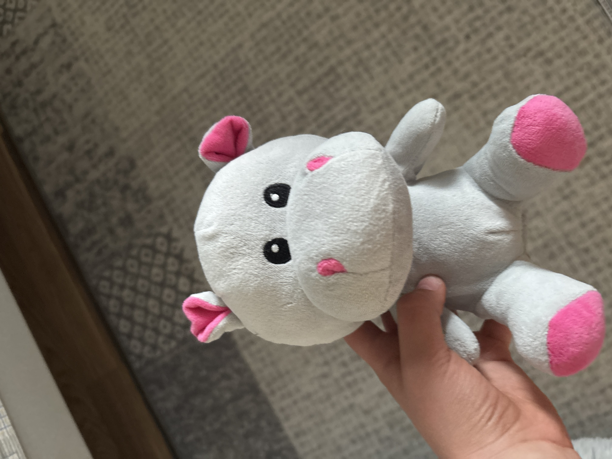 A grey plush hippopotamus toy with pink accents is held by a person's hand against a carpeted floor and wooden furniture. The hippopotamus has pink inner ears, a pink nose, and pink soles on its feet. Its eyes are black and round. The background shows a textured carpet with a geometric pattern and the edge of a wooden surface, possibly a bed frame or dresser. The lighting suggests an indoor environment. The location is Unknown, Unknown.