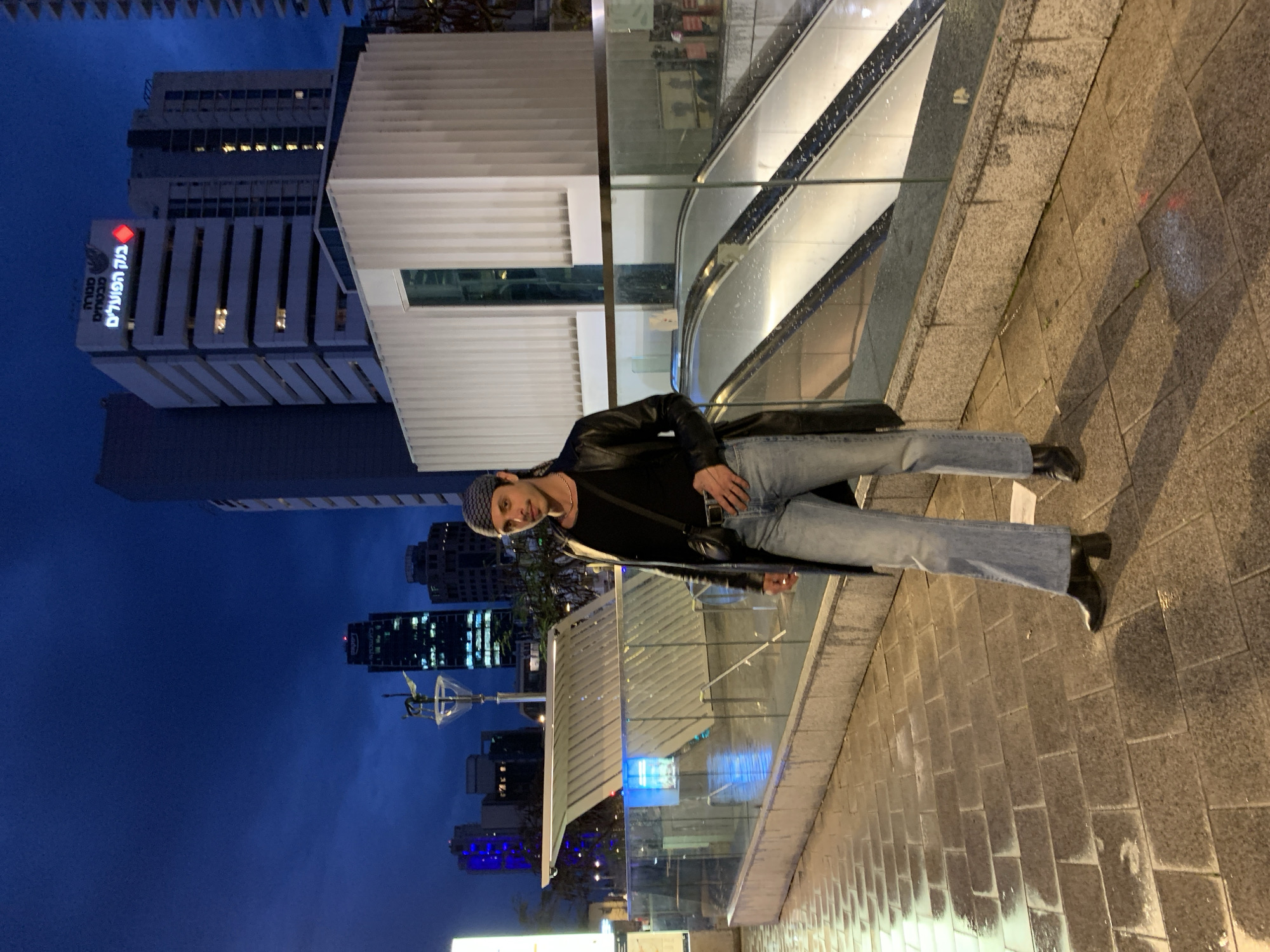 A man wearing a dark-colored jacket, jeans, and a patterned cap poses for a photo on a sidewalk in Tel Aviv, Israel. The background includes modern buildings, one with Hebrew text that reads "בנק הפועלים" (Bank Hapoalim). An escalator is visible behind the man. The time appears to be evening or night.
