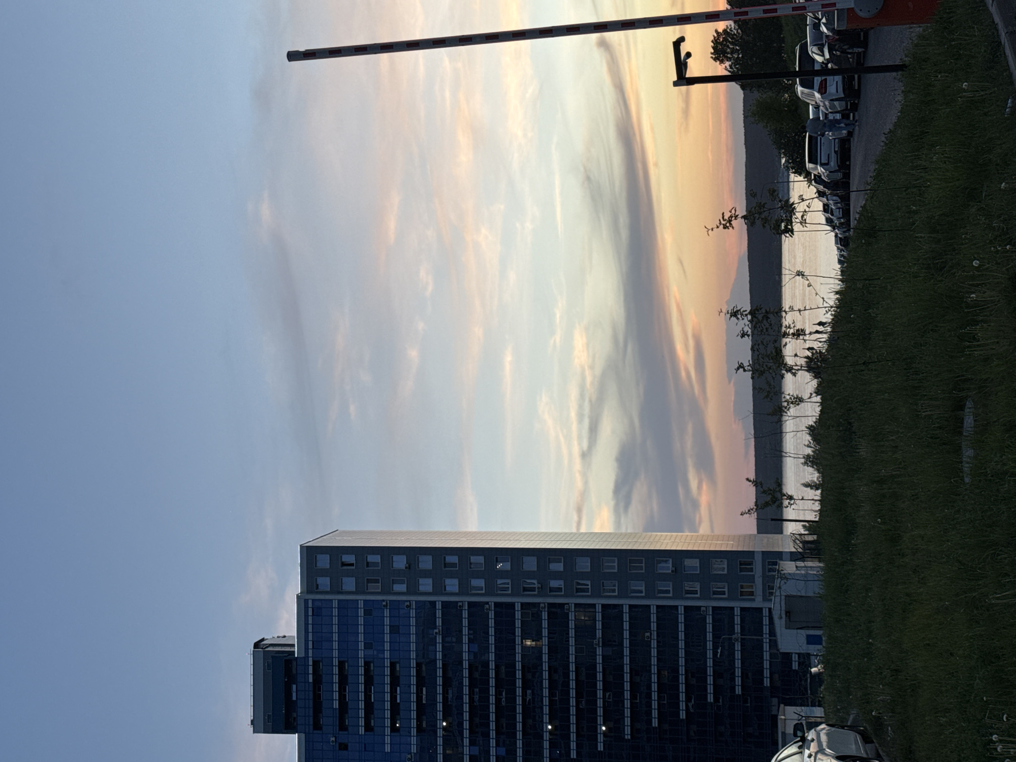 The image shows a view from ground level with a modern high-rise building dominating the foreground. The building features a facade primarily composed of glass windows, indicating a likely office or residential structure. Above the building, the sky is visible, exhibiting a gradient of light blue to orange hues, potentially indicating sunrise or sunset. A striped barrier arm extends across the upper portion of the image, suggesting a controlled access point. Beyond the barrier, a row of vehicles is parked alongside a patch of green vegetation. In the distance, a body of water and land can be seen. The overall impression is an urban setting with a mix of natural and architectural elements during a transition period of daylight.