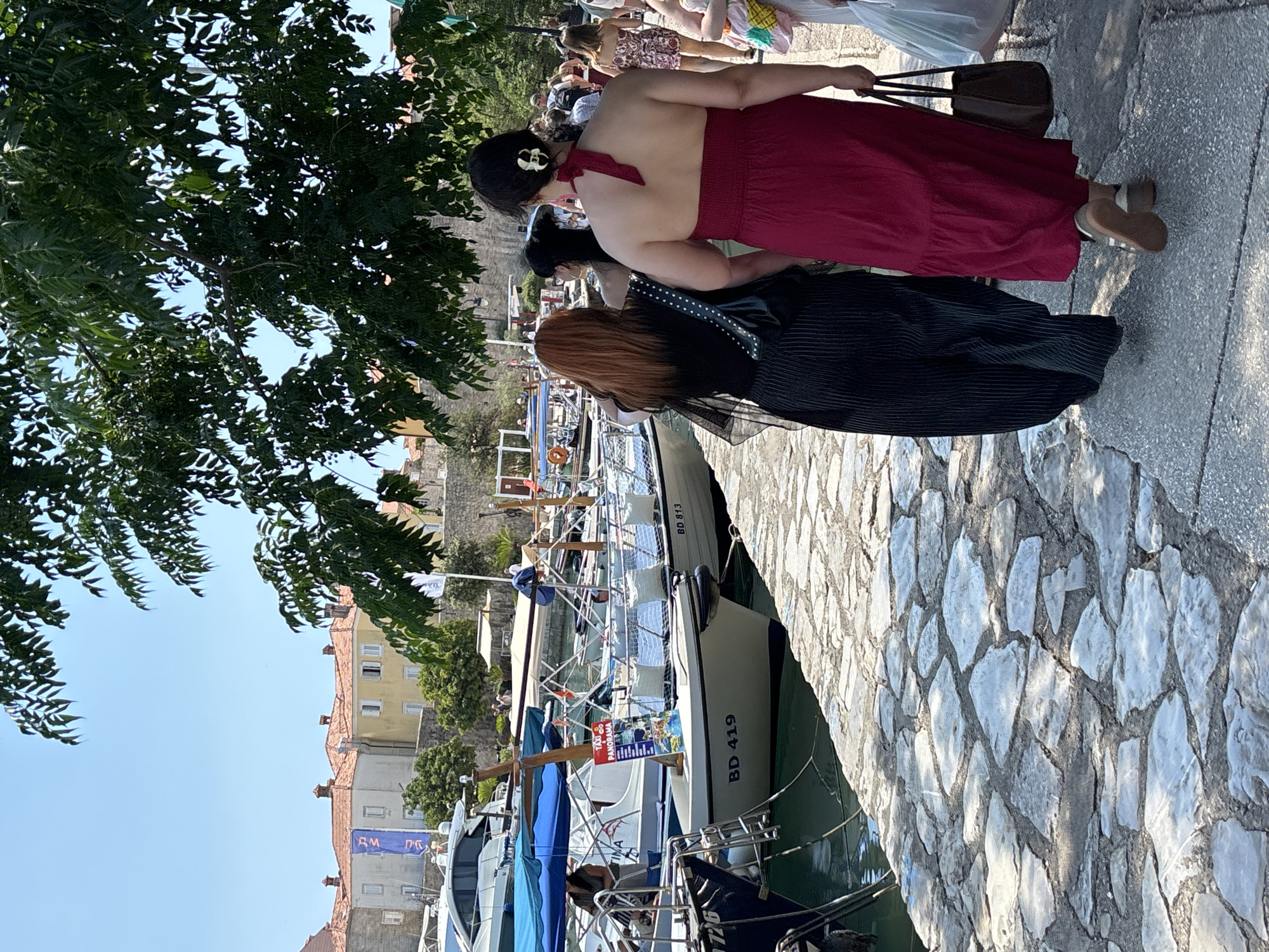 The image depicts a sunny day in Budva, Montenegro, focusing on a stone pathway adjacent to a harbor filled with boats. In the foreground, two women walk away from the camera on the pathway. The woman on the left has red hair and wears a black, sheer, long-sleeved top and striped pants. She carries a black purse. The woman on the right is wearing a strapless red dress and carries a brown tote bag. Behind them, several boats are docked. One boat, with the registration "BD 419" visible, has a "TAXI PANORAMA" sign on it. Other boats display various blue and white flags. In the background, traditional Mediterranean buildings with orange tiled roofs line the waterfront, and a stone wall runs along the embankment. Lush green trees canopy the scene, casting shadows on the pathway and water. Other pedestrians are visible in the distance.