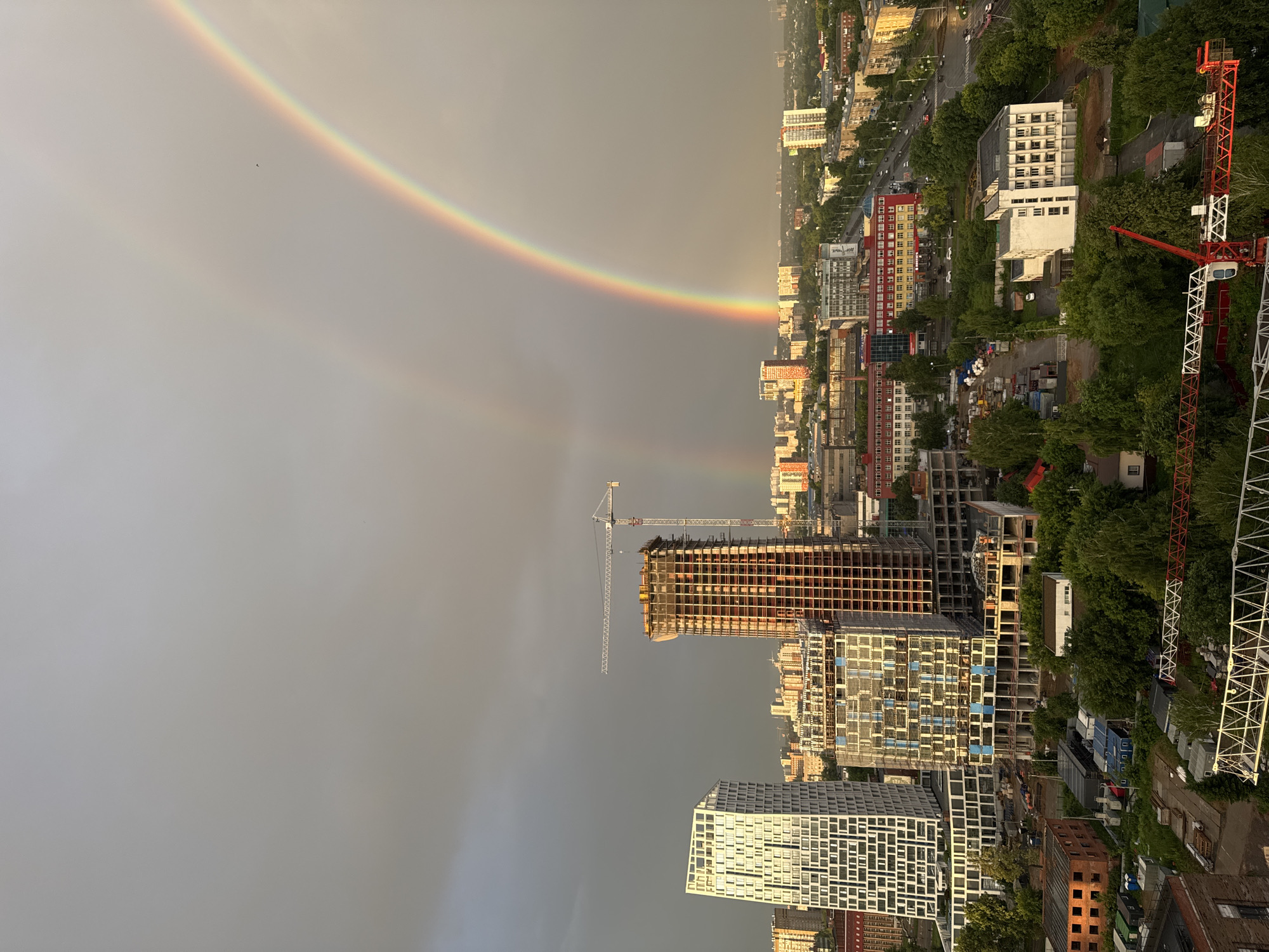 The image depicts a cityscape, possibly Izhevsk, Russia, with a prominent double rainbow arching across a cloudy sky. In the foreground and midground, construction sites are visible, featuring several tall buildings under various stages of completion. Notably, there are cranes, including a large tower crane. The buildings range in style, from modern, geometrically patterned structures to older, more conventional architecture. A street with some vehicle traffic runs through the scene. Green trees and foliage are interspersed throughout the urban landscape. The overall impression is of a city undergoing development under a dramatic sky.