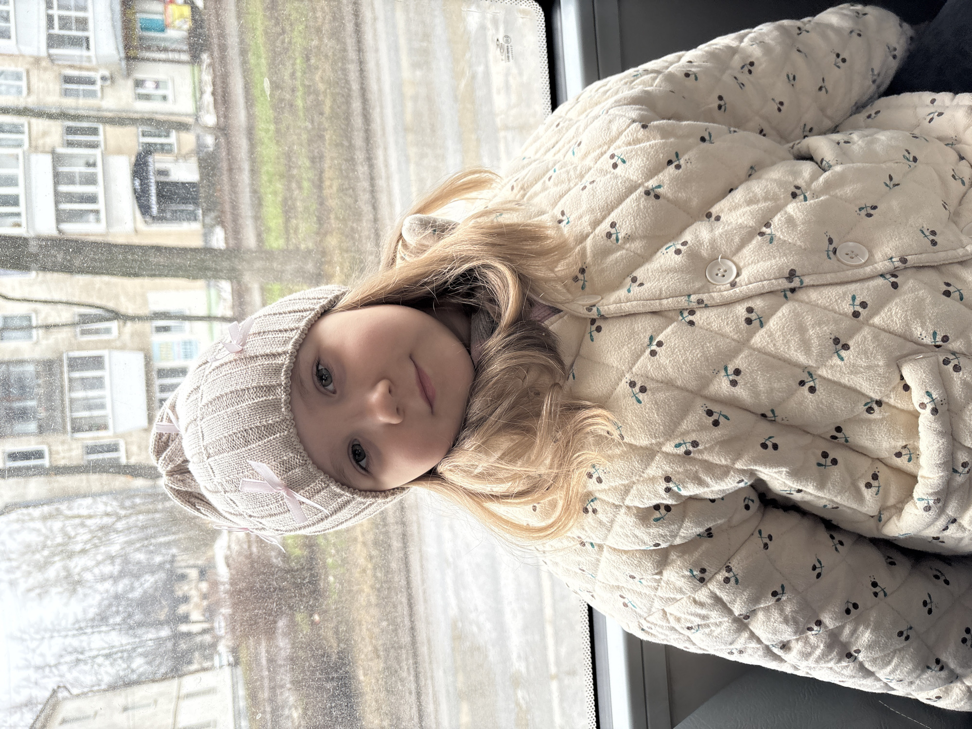 A young girl sits on a gray seat inside a vehicle, possibly a bus or tram, as evidenced by the view through the window. The girl is wearing a beige knit hat with a bow and a quilted jacket with a cherry pattern. She has blonde hair and blue eyes, and she is looking directly at the camera. The background shows an urban environment with apartment buildings and bare trees, suggesting it might be winter or early spring. Based on the query, the location is likely the city of Chaikovsky, Russia. The window is somewhat smudged, indicating recent precipitation or lack of cleaning.