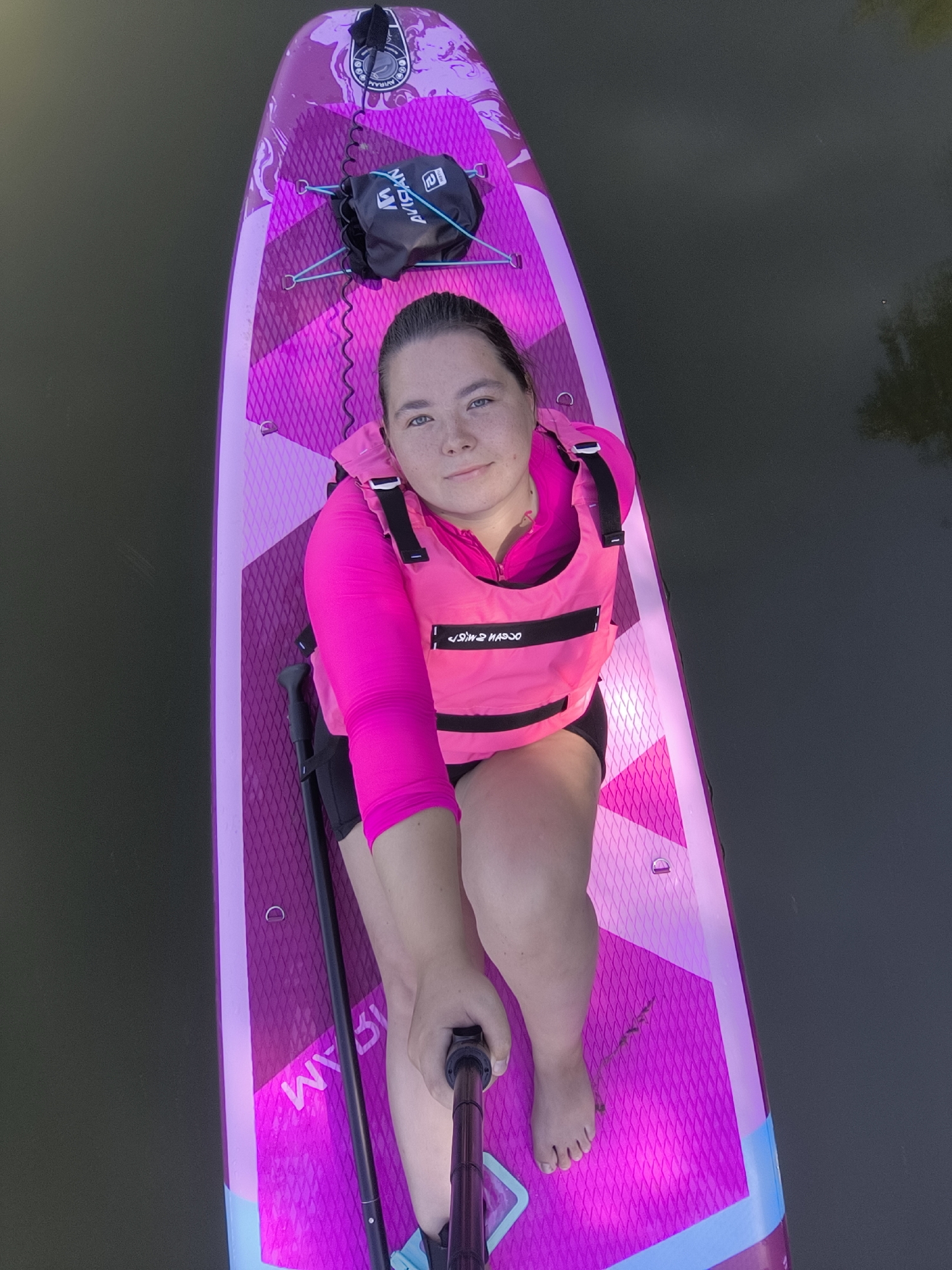 A woman is sitting on a bright pink paddleboard, looking up at the camera. She is wearing a bright pink life vest over a long-sleeved pink shirt, and black shorts. Her feet are bare. She is holding a paddle. The paddleboard is on dark water. There is a black bag with blue straps attached to the front of the paddleboard. The image appears to have been taken from a slightly elevated perspective. It is not possible to determine the specific location within Kaliningrad, Russia based on the visual information provided.