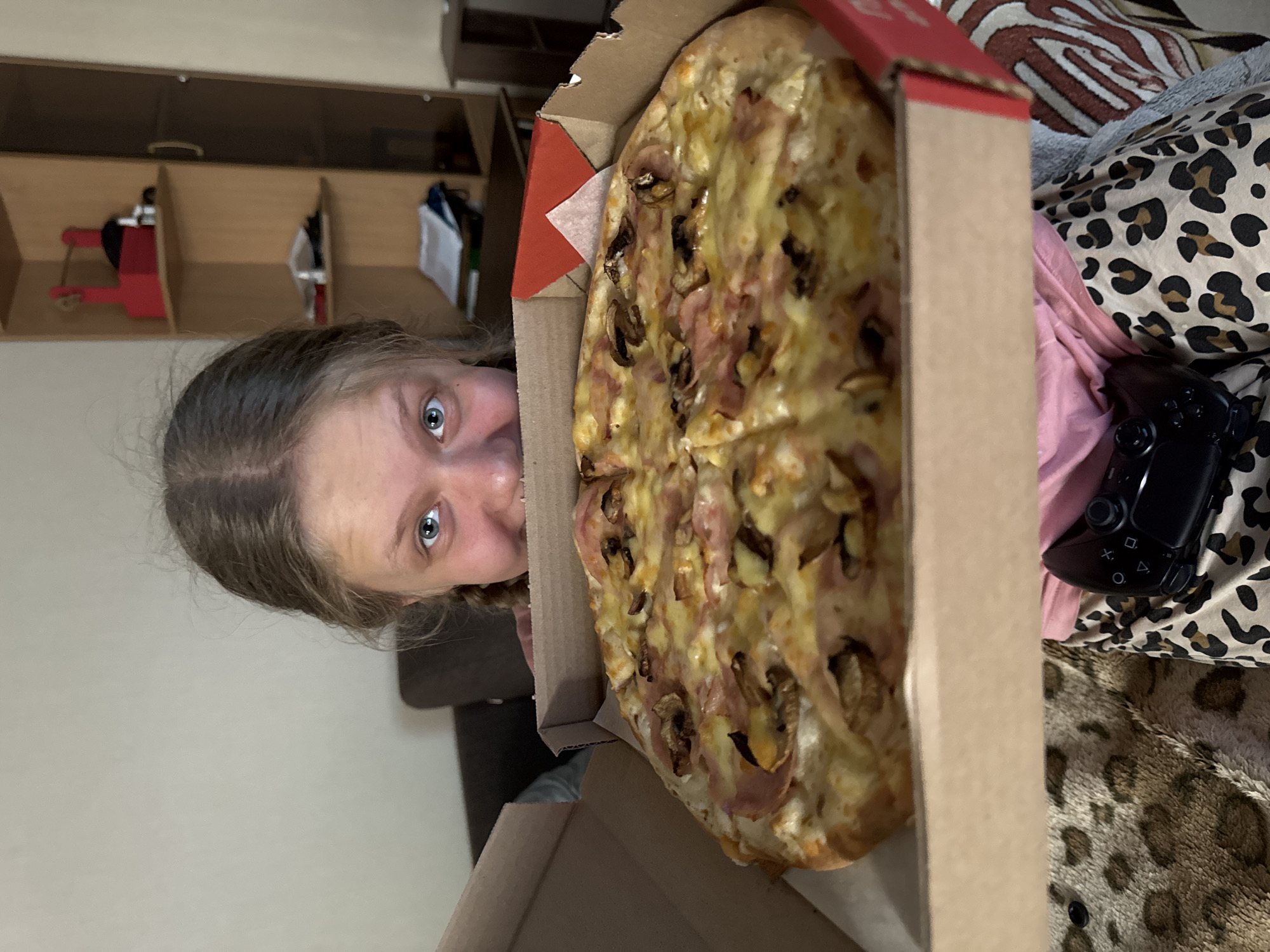 A person with light brown hair is looking at a pizza in a cardboard box. The pizza is topped with cheese, mushrooms, and ham. A black Playstation controller is on the right side of the image. The background includes a cabinet and a leopard-print pillow. The image was taken in an apartment in Chaikovsky, Russia.