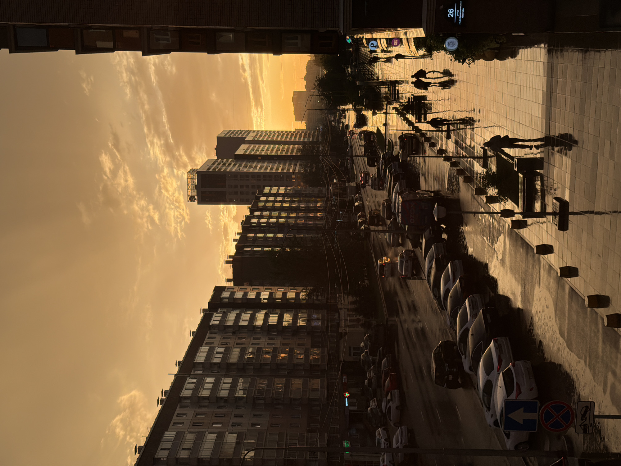 The image depicts a street scene in Krasnoyarsk, Russia, during sunset. The sky is a vibrant orange and yellow, with clouds casting long shadows. Buildings, some residential and others modern high-rises, line both sides of the street. The street and sidewalk are wet, indicating recent rain, and reflect the warm light of the setting sun. Cars are parked along the street and others are driving, their headlights creating streaks of light. Several people are walking on the sidewalk, their silhouettes dark against the bright reflections. One person is walking alone, while a group of three is walking together further down the sidewalk. Traffic signs are visible, including a parking sign and a sign indicating a no parking zone. A commercial sign for "OZON" is visible on a van. The overall atmosphere is one of urban life transitioning into evening under a dramatic sky.