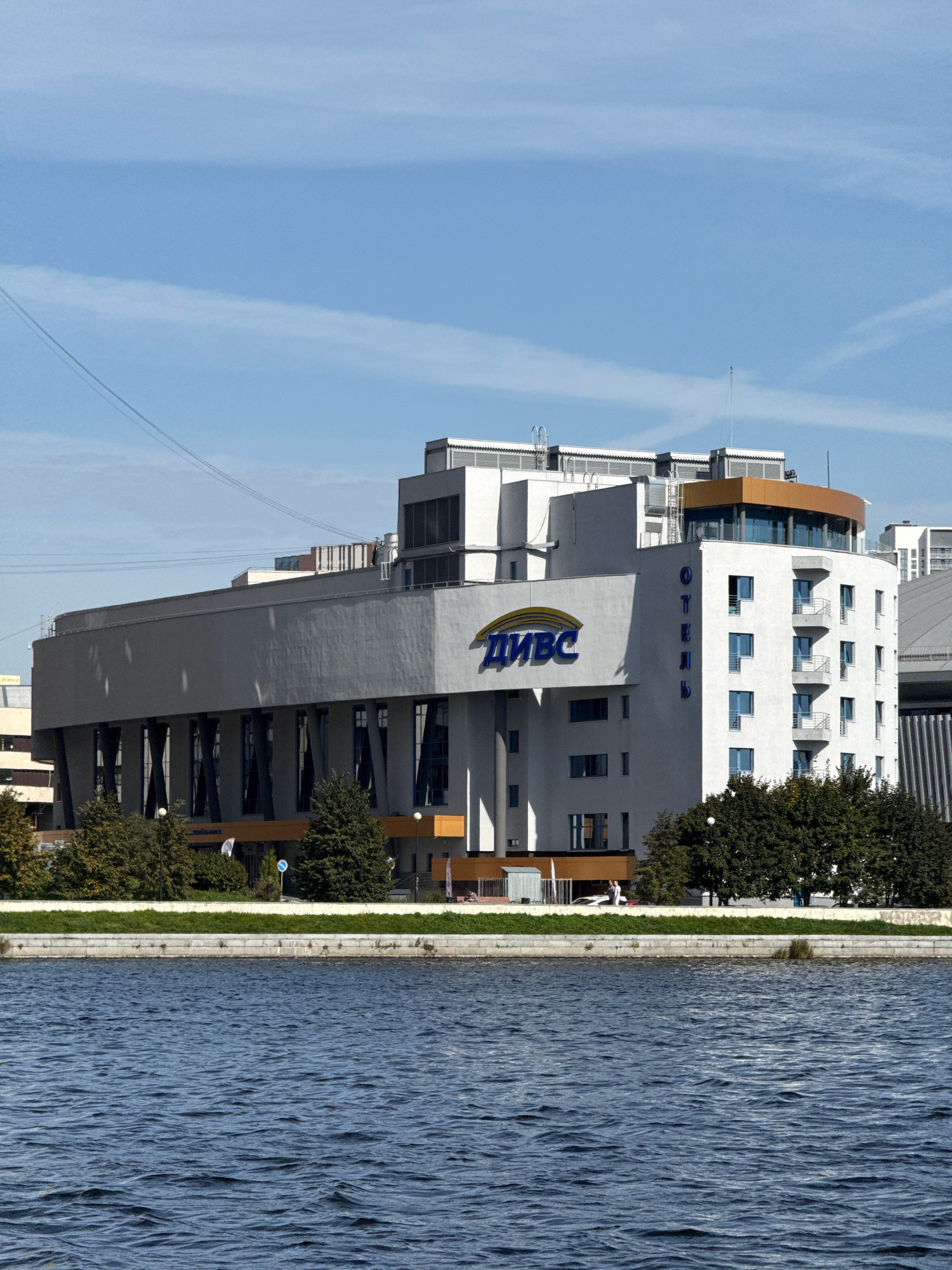 This is a medium-shot, eye-level, full-frame, exterior photograph of the DIVS (Palace of Sports) located in Yekaterinburg, Russia, on a clear day. The building is a large, multi-story structure with a modern architectural design, situated on the bank of the Iset River. The river occupies the lower portion of the frame, its dark blue water disturbed by ripples.
The DIVS building is primarily white and gray, with contrasting elements of orange and blue. A prominent blue logo with the Cyrillic letters "ДИВС" is visible on the left side of the building, arching over a dark gray facade punctuated by tall, narrow windows. To the right, a section of the building is white and features a series of balconies with gray railings. Vertical blue Cyrillic lettering spelling "ОТЕЛЬ" (Hotel) is also visible on this white section. The upper part of the building has several levels with windows and some exposed mechanical equipment.
Trees with lush green foliage are planted in front of the building, forming a barrier between the structure and the river. A low, gray stone wall separates the grass from the riverbank, and a strip of neatly trimmed green grass runs along the top of the wall.
In the background, parts of other buildings and structures are visible, including a lighter-colored building with windows and a tall, thin antenna or flagpole. The sky is a bright, clear blue with faint, wispy white clouds.
There are no visible individuals or animals in the immediate foreground or on the building's facade. A few indistinct figures can be seen on the embankment in the distance. The overall scene depicts a clean, urban waterfront environment.