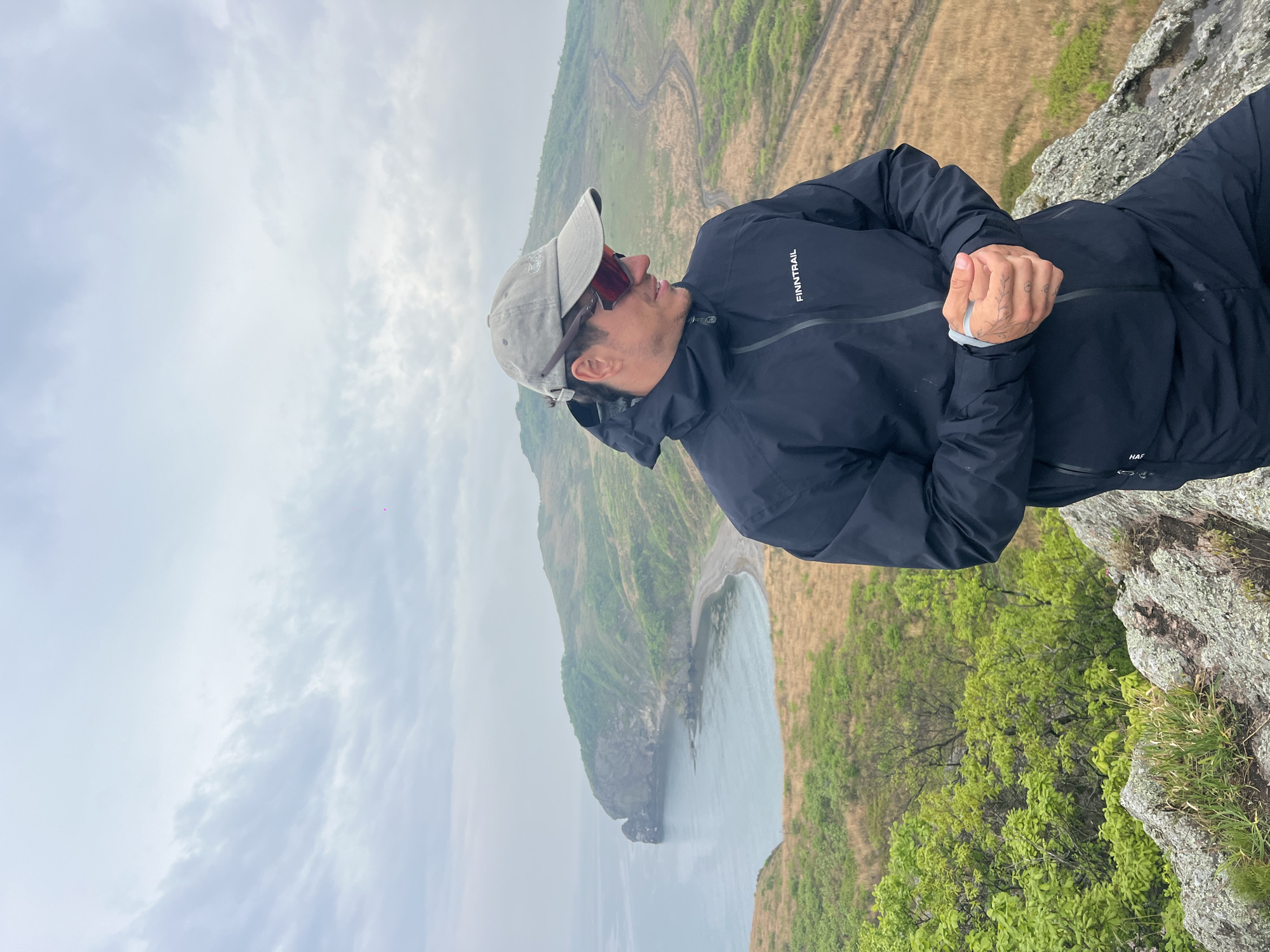 A man in a dark jacket, light-colored cap, and sunglasses is standing on a rocky outcrop. He is looking downwards and slightly to his left. The landscape behind him features a hillside with a mix of green vegetation and brown, grassy areas. In the distance, there is a body of water and a cloudy sky. The location is Gvozdevo, Russia.