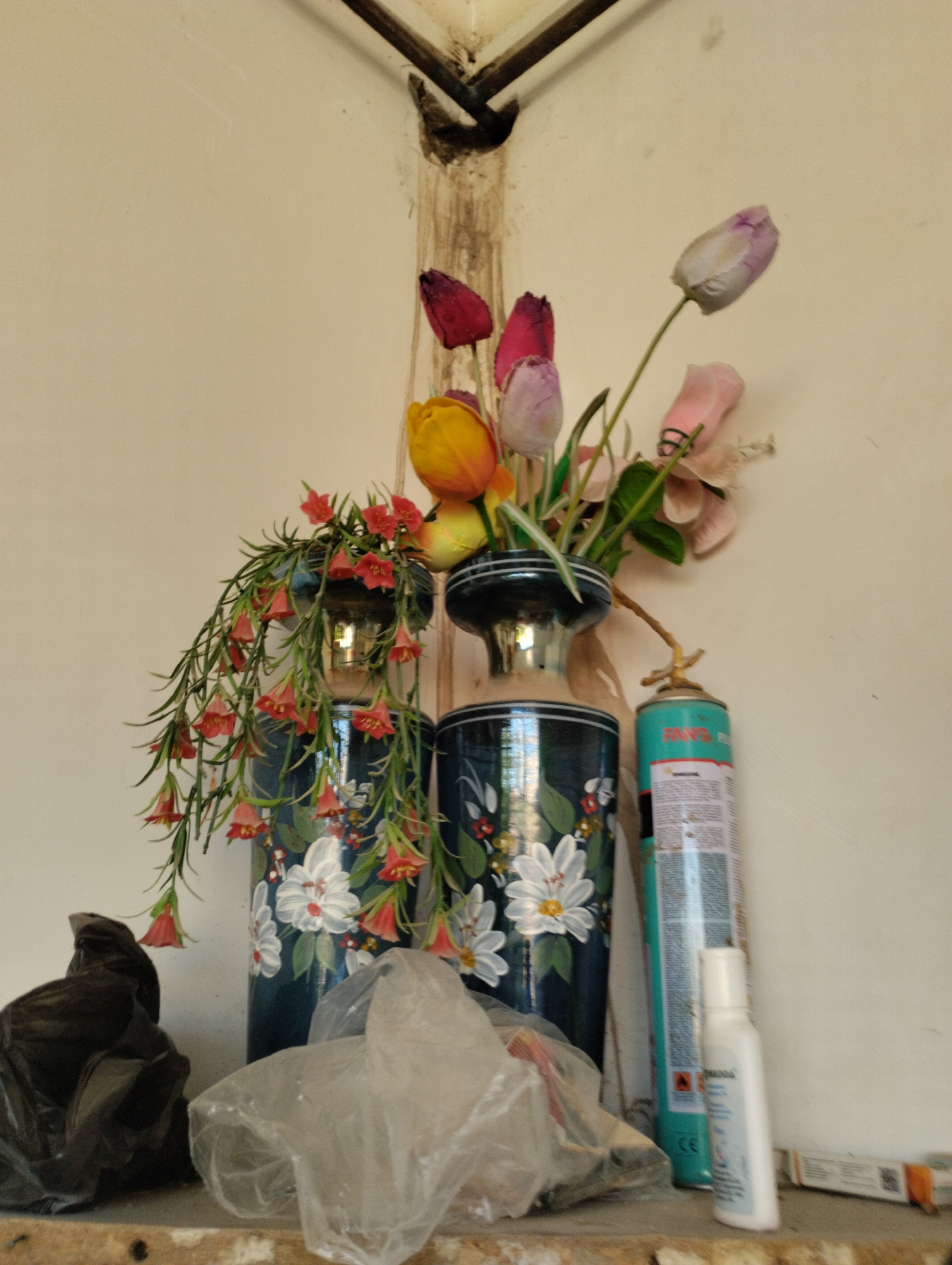 In a dimly lit corner with a light-colored wall, a shelf holds several items. Two dark blue vases with white floral designs are prominent. The vase on the left has a cascade of small red bell-shaped flowers and green foliage spilling out. The vase on the right contains a bouquet of artificial tulips in shades of red, yellow, pink, and purple. To the right of the vases, a teal spray can with white lettering stands upright, and a small white bottle is positioned in front of it. In the foreground, on the shelf, are two crumpled plastic bags, one black and one translucent. The shelf itself appears to be made of unfinished wood. A dark metal pipe is visible against the wall in the upper portion of the image.