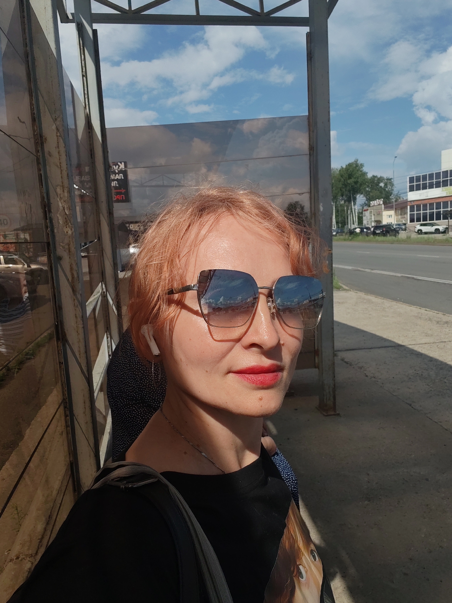 The image shows a woman with short, reddish-blonde hair, wearing large, reflective sunglasses and red lipstick. She is standing outdoors, possibly at a bus stop, with a bus stop shelter structure to her left. Reflected in her sunglasses are a blue sky with clouds and some buildings. Behind her, to the right, is a road with some cars and buildings visible in the distance. A portion of a building with a grid-like facade is also visible. The overall impression is of a sunny day in an urban or semi-urban environment. It is not possible to definitively confirm the location as Chaikovsky, Russia, based solely on the visual information provided in the image.