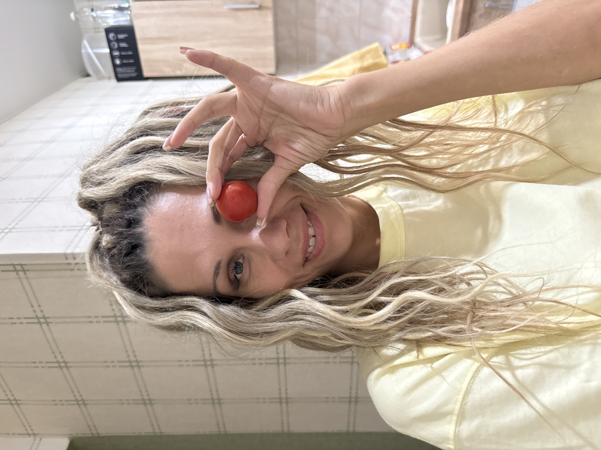 A woman is holding a cherry tomato in front of her right eye, obscuring it. She is looking at the camera and smiling. She has long, blonde, wavy hair and is wearing a light yellow t-shirt. The background appears to be a kitchen with light-colored tiled walls and wooden cabinets. The image is a medium shot, focusing on the woman's face and upper body. The specific location of Rusanovo, Russia is not discernible from the visual information in the image.