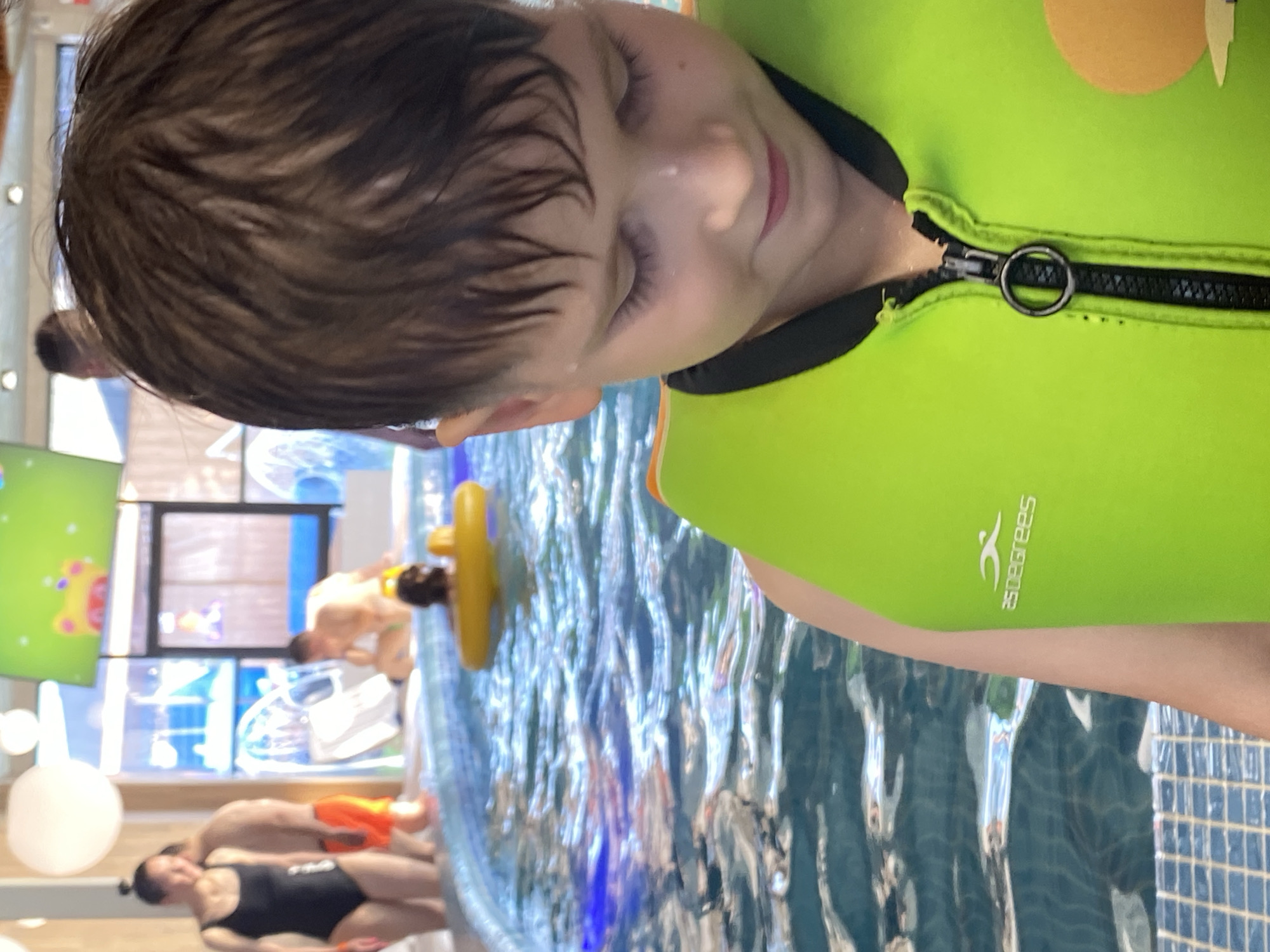 The image shows a young boy wearing a bright green life vest in what appears to be an indoor water park. The boy is positioned in the foreground, looking down with a slight smile. The water in the pool is blue and reflects light, with ripples visible on the surface. In the background, other people are visible, some swimming or wading in the water. One person is wearing a yellow float. The environment suggests an indoor recreational facility with bright lighting and possibly some form of entertainment displayed on a screen overhead. The tiled edge of the pool is visible in the lower part of the frame. There is no information about the specific location in Votkinsk, Russia, within the image itself.