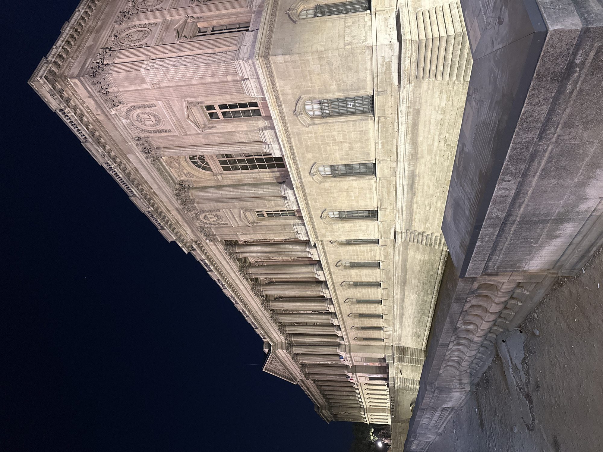 An upward-angled, low-light shot captures the ornate facade of a grand, classical-style stone building at night. The structure is illuminated by artificial lights, giving its pale stone a warm, yellowish-beige glow against a dark, starless navy-blue sky.
The building features multiple levels with numerous windows, many of which are rectangular with grates, while some are arched. A prominent architectural feature is a long colonnade of classical columns, stretching along the building's length. Above these, the facade is richly decorated with intricate carvings, including detailed cornices, friezes with decorative patterns, and circular motifs resembling wreaths or medallions. The style suggests a significant public building, possibly in Paris.
In the immediate foreground, a large, dark stone wall or plinth partially obscures the lower part of the building. This structure has a rugged, possibly weathered texture. On a darker, flat section of this foreground element, faint, unreadable text or etchings are visible, appearing as rows of small characters or illustrations. No people or specific activities are visible, emphasizing the monumental architecture. The overall impression is one of historical grandeur seen under a clear night sky.