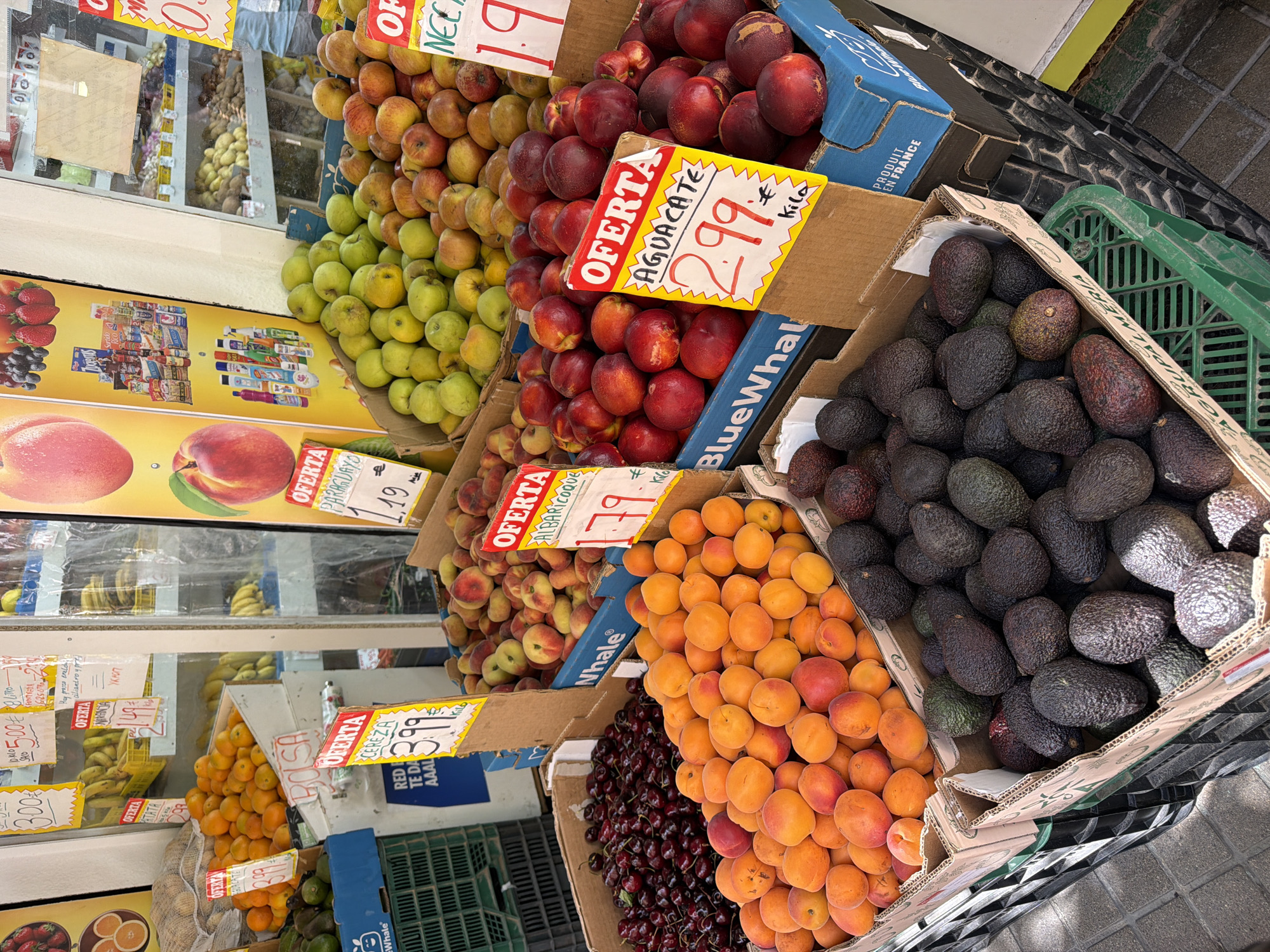The image shows a fruit stand in what appears to be a market. The stand is filled with various fruits, including apricots, cherries, avocados, peaches, and nectarines, all displayed in cardboard boxes and plastic crates. Some of the fruits have price tags attached, with prices like "3.99" and "2.99" visible. In the background, shelves are stocked with more fruits and packaged goods. The ground is paved with grey tiles. There are no people clearly visible in the image, but the presence of the market setting implies a public space. The lighting suggests it is daytime.