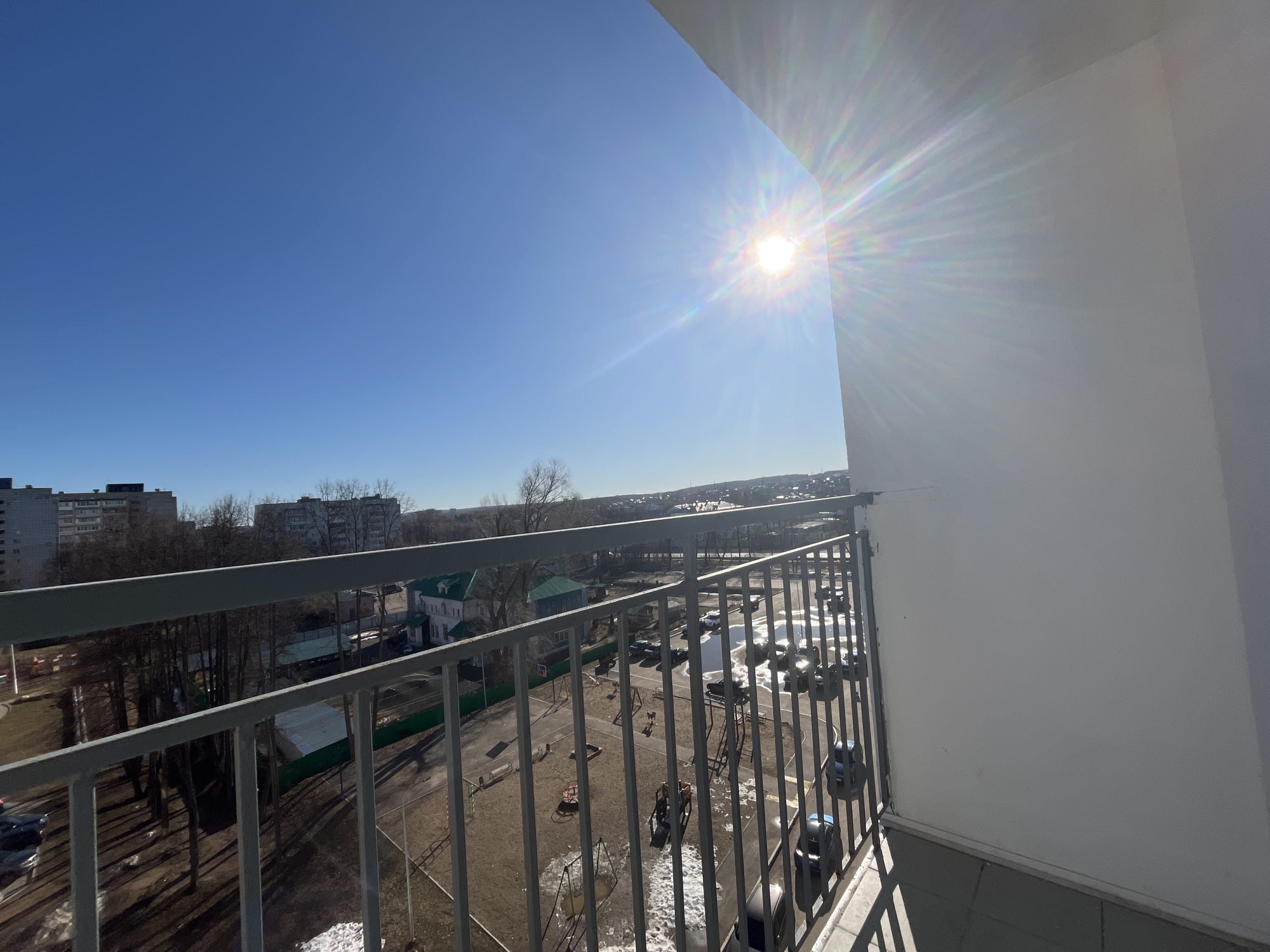 The image is a high-angle view from a balcony, likely in an apartment building, located in Chaikovsky, Russia. The sky is a clear blue, and the sun is high and bright, causing lens flare in the image. The balcony has a gray metal railing. Below, there is a playground with various play structures, including swings and climbing frames. Cars are parked nearby. Some patches of snow are still visible on the ground. In the background, there are residential buildings and trees, indicating an urban or suburban area.