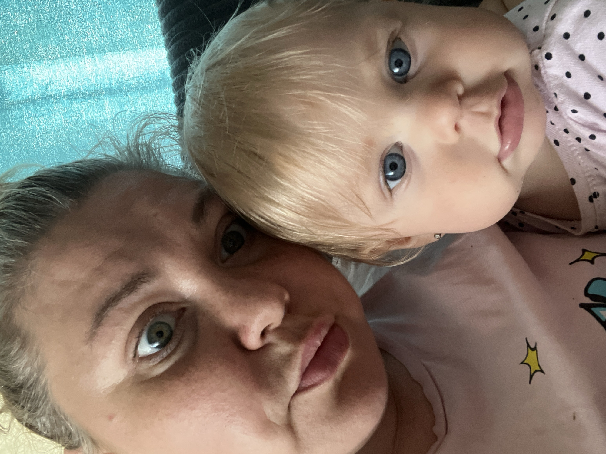 This is a selfie of two people, a woman and a baby, likely taken indoors. The woman is in the foreground on the left, with her face angled towards the camera. She has light-colored hair pulled back and her eyes are wide open, looking directly at the camera. Her lips are slightly pursed. She is wearing a light-colored t-shirt with a yellow star design on it. To the right of the woman is a baby, facing forward. The baby has fair hair and large blue eyes. The baby is wearing a pink and black polka dot shirt. The background appears to be a turquoise textured curtain. The image does not contain any information about the location in the city of Фоки, Russia.