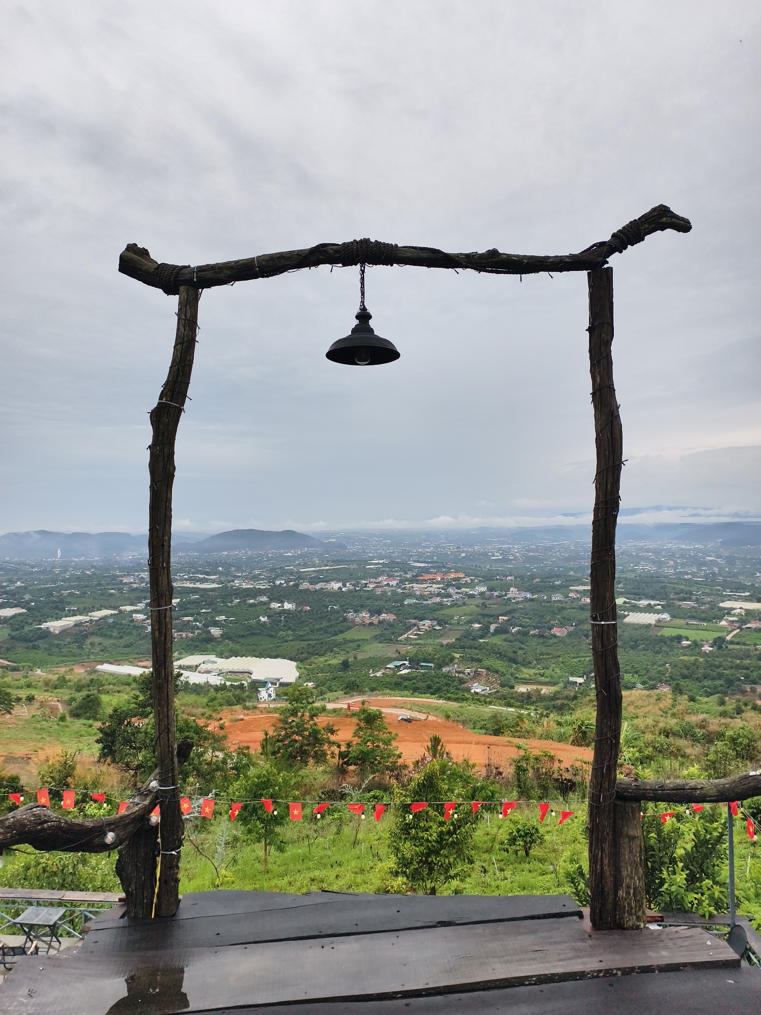 A wide, eye-level shot depicts a view from a rustic wooden platform overlooking a cityscape. The platform is constructed from dark-stained planks, appearing slightly wet. Two rough-hewn wooden posts flank the view, connected by a horizontal beam at the top, from which hangs a single black lamp.
In the background, a landscape unfolds with a mix of green foliage and scattered buildings. A patch of red earth is visible on the hillside. Red flags with yellow stars are strung along the lower wooden structure.
In the far distance, hazy mountains can be seen against a cloudy sky. The overall impression is of a scenic overlook in Trung Hà, Vietnam, likely designed to provide a viewpoint.