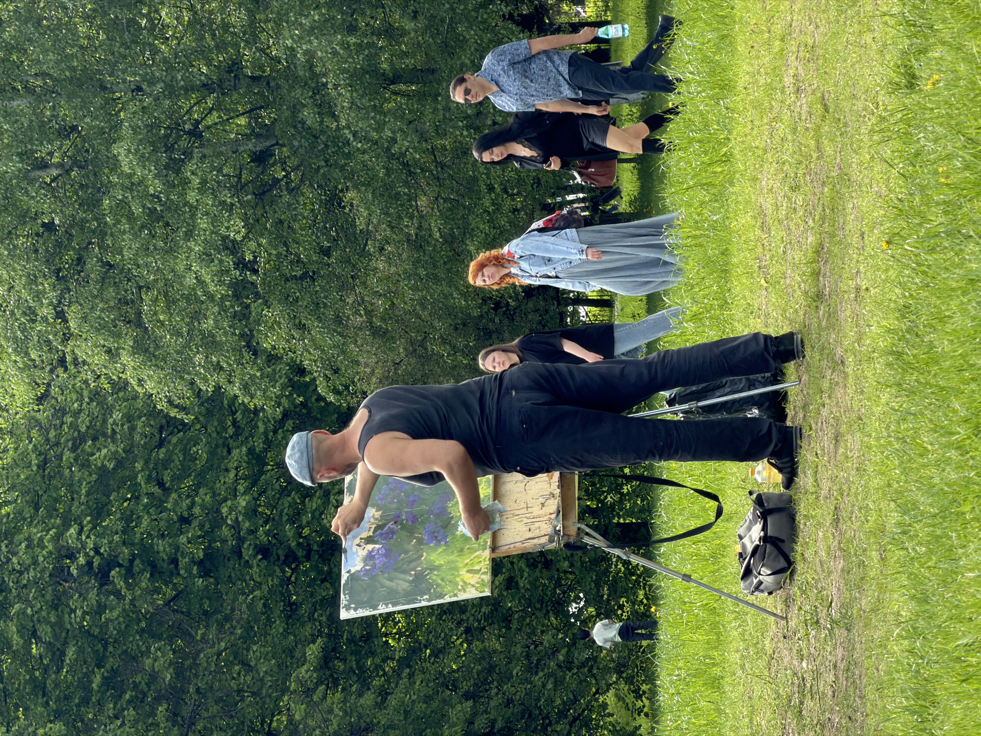 An artist is painting a landscape en plein air in Moscow, Russia. The painter, wearing a tank top and a light blue hat, is standing in front of an easel. The painting displays an image of green grass and purple flowers. Behind him are four people, including one woman with red hair wearing a blue denim dress. The artist's black bag and easel stand on a grass field. Trees stand in the background.
