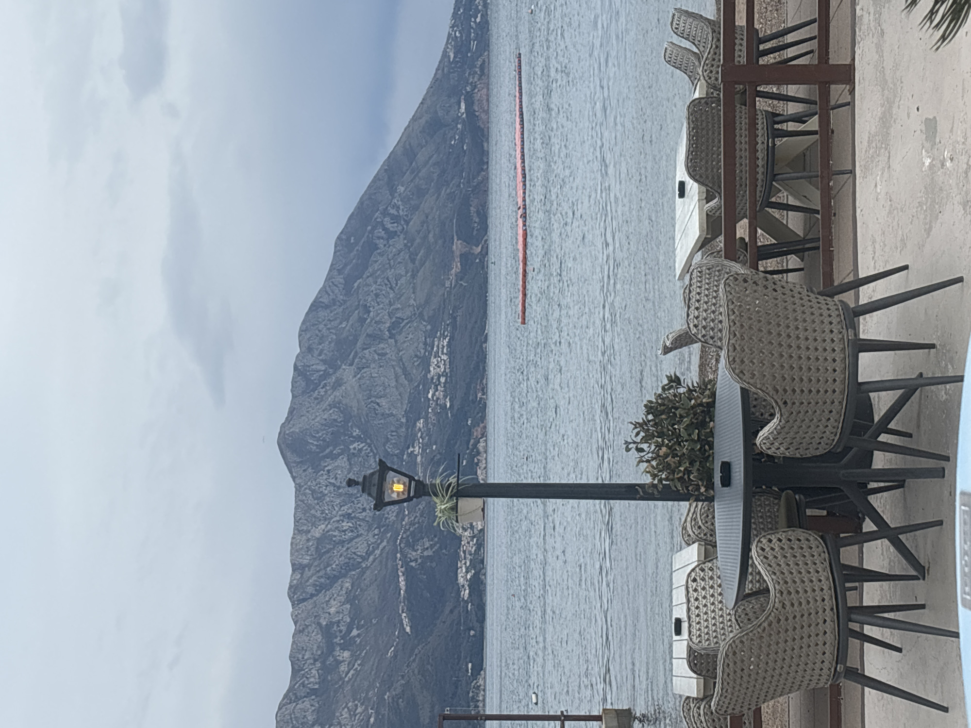 The image displays a picturesque coastal or lakeside scene, captured from what appears to be an outdoor cafe or restaurant patio. The original image is oriented sideways, but when viewed correctly (landscape), it shows a body of water with mountains in the background and outdoor seating in the foreground.
In the foreground, on the right side of the frame, there is an outdoor seating area. Several tables and chairs are visible. The chairs are made of a woven material, likely rattan or wicker, in a light brown or beige color, with dark metal legs. The tables are dark gray or black. To the left of the seating area, a black street lamp post stands tall, with an illuminated warm yellow light bulb inside its lantern-style fixture. A small plant is attached to the lamp post. Another smaller, bushy green plant is situated between two of the tables. The ground of the patio appears to be light-colored concrete or paving.
The midground is dominated by a large body of water, likely a sea or a large lake, extending from the patio edge towards the distant mountains. The water surface is slightly choppy, reflecting the overcast sky. A prominent feature in the water is a long, bright orange floating barrier or walkway, stretching horizontally across a significant portion of the frame. It appears to be made up of numerous interconnected segments.
In the background, majestic, rugged mountains rise steeply from the water's edge. The mountains are predominantly dark gray, indicating a rocky terrain, with some patches of lighter color that might be small villages or settlements nestled along their base. The sky above the mountains is overcast, filled with light gray clouds, suggesting a cloudy day.
The overall lighting is soft and diffused, consistent with an overcast day. Despite the cloudy conditions, the light from the street lamp suggests it might be late afternoon or early evening, or simply turned on during the day. There are no visible people or discernible text in the image. The scene conveys a calm and serene atmosphere, inviting contemplation of the natural landscape from a comfortable vantage point.
