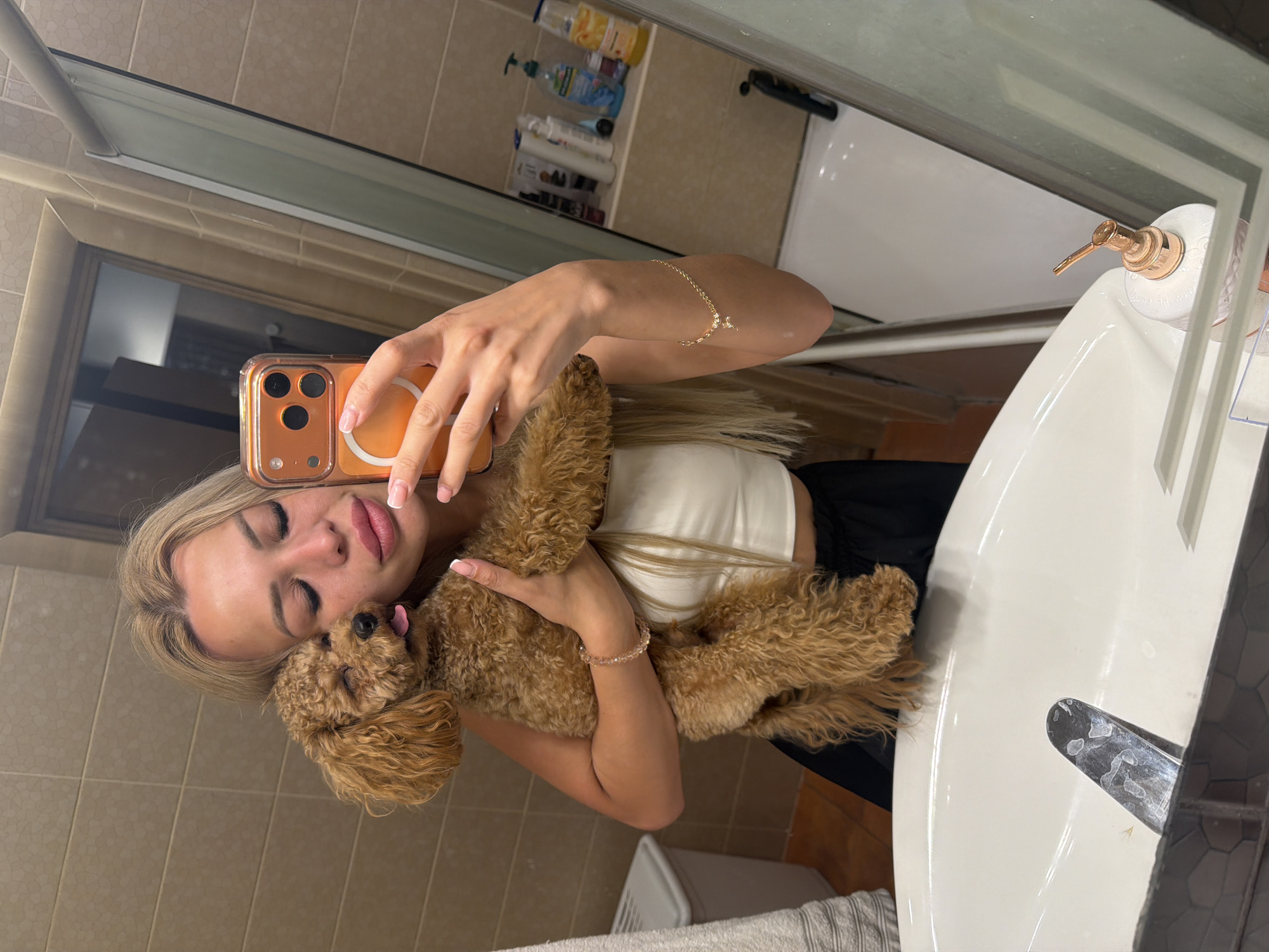This is a vertical, slightly off-center selfie taken in a bathroom. The main subject is a young woman with blonde hair, wearing a white tank top and black bottoms. She is holding a small, curly-haired brown dog, which is nuzzling her face. The woman is looking at her phone, which is out of frame, and appears to be taking the photo. Her eyes are closed, and she has a soft smile, conveying a sense of affection towards the dog. She is wearing a delicate bracelet on her wrist.
The background shows a tiled wall and a bathroom sink with a rose gold soap dispenser. In the reflection of a mirror above the sink, several bottles of toiletries can be seen on a shelf. The overall lighting suggests it might be daytime, and the setting appears to be a residential bathroom. The mood is intimate and peaceful, highlighting the bond between the woman and her pet. There is no visible text in the image. The location context is Kyiv, Ukraine.