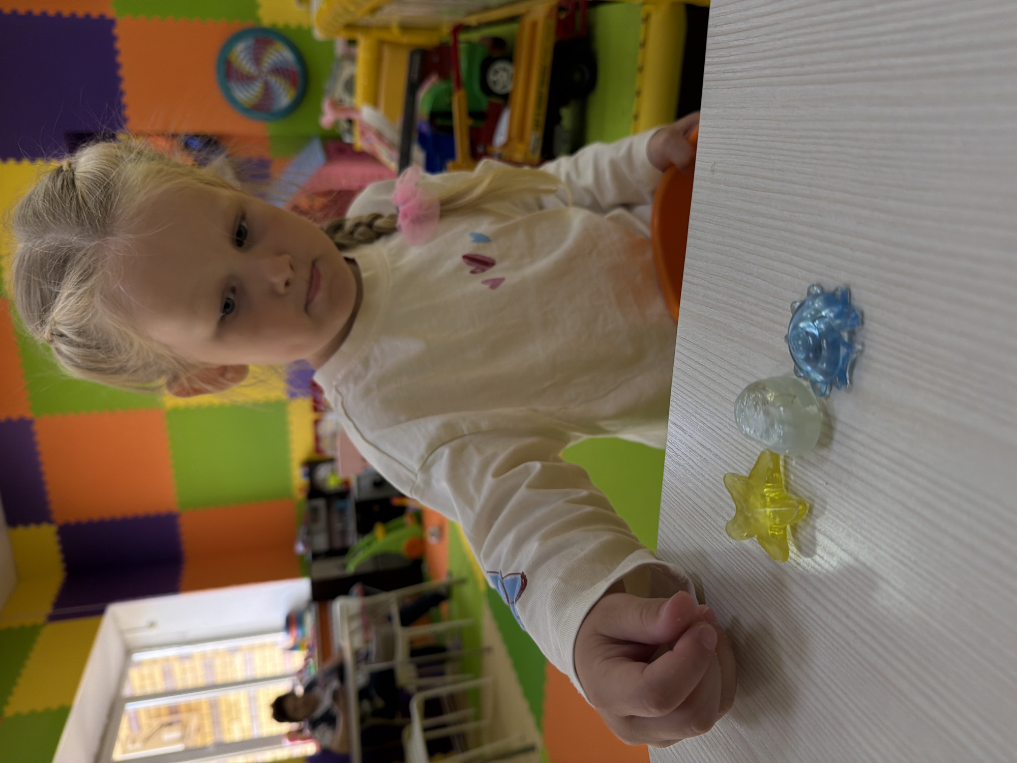 A young Caucasian girl with blonde hair in two braids, adorned with a pink scrunchie, is looking down at a wooden table. She is wearing a long-sleeved cream shirt with small embroidered hearts on the sleeve. On the table in front of her are three translucent, jewel-like objects: a yellow star, a clear oval shape, and a blue star-shaped object with a ridged pattern. The girl's left hand is near the objects. The background is a brightly colored room with a wall covered in foam squares in orange, green, yellow, and purple. A window with blinds is visible on the left, and other children and adults are in the blurred background. A yellow toy structure with toy vehicles is also visible to the right. The scene appears to be a classroom or play area. This image was taken in Kamyshin, Russia.