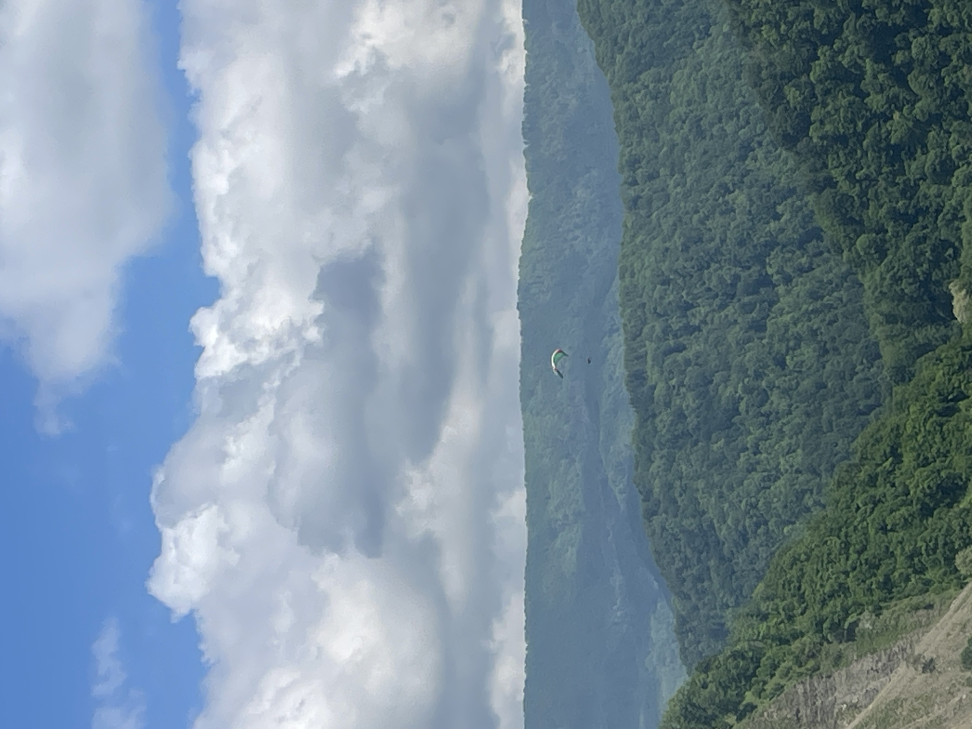 The image shows a mountainous landscape under a partly cloudy sky. A paraglider is visible in the middle ground, positioned between the mountain slope covered with lush green forest and the clouds. The sky transitions from blue to gray with the presence of white and gray clouds. The image does not provide enough information to confirm the location is Baranovka, Russia. The environment appears to be a natural setting, with the prominent features being the mountains, forest, and sky. There are no visible interactions or other objects, subjects, or people besides the paraglider.