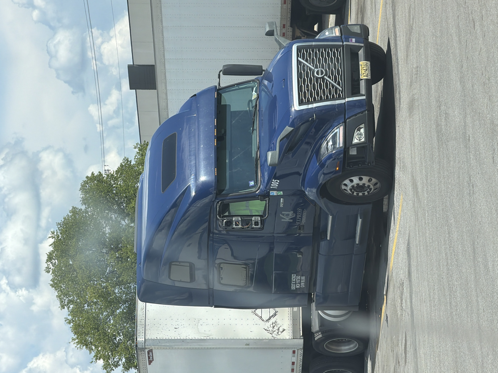 A dark blue Volvo semi-truck, labeled "K&L EXPRESS LOGISTICS" and numbered "1005", is parked in a paved lot in Lebanon, United States of America. The truck has a white trailer attached and is positioned in front of a light-colored building with a dark rectangular panel. The sky is partly cloudy, with green trees visible in the background. A yellow line runs across the pavement in front of the truck. The truck's license plate reads "AZ 4760".