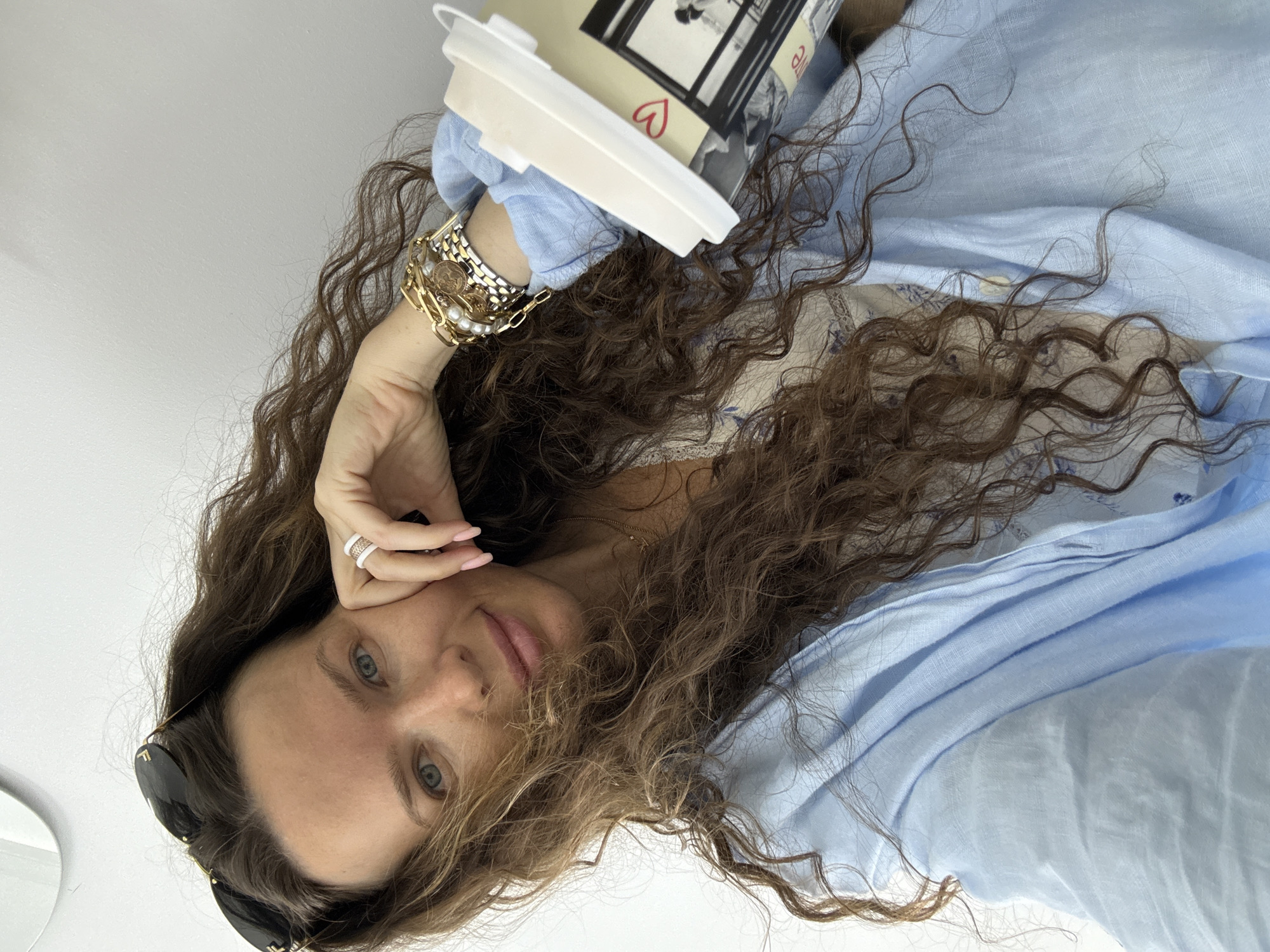 A medium shot photo shows a woman with long, curly brown hair and blue eyes. She is wearing a light blue button-down shirt. She is also wearing sunglasses on top of her head, multiple bracelets on her left wrist, and a ring on her right index finger. The woman is holding a white disposable coffee cup in her right hand. The background is a white wall, and a sliver of a round mirror is visible in the upper left corner. The image has a shallow depth of field, with the woman's face and upper body in sharp focus, and the background blurred. The scene is lit with natural light, coming from the right side of the image. The overall impression is that of a casual, candid moment. It is not possible to determine the location in Moscow, Russia based on the visual information provided.