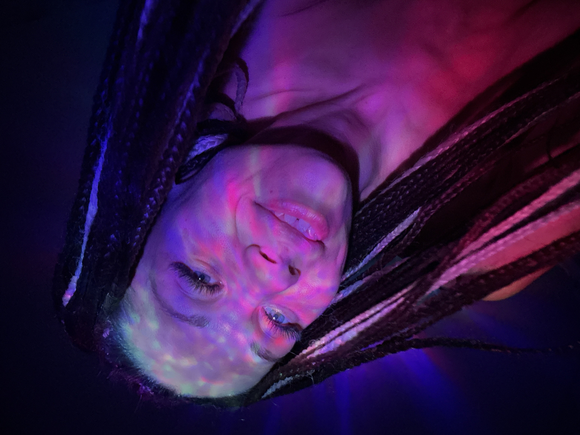 A close-up, tilted photograph depicts a person illuminated by pink and blue light, possibly from a disco ball. The person has fair skin, blue eyes, and dark hair styled in braids with occasional white or silver strands. Their expression appears to be a slight smile. The background is dark, emphasizing the colorful lighting. The photograph appears to be taken indoors. The location (Rusanovo, Russia) is not apparent within the image itself.