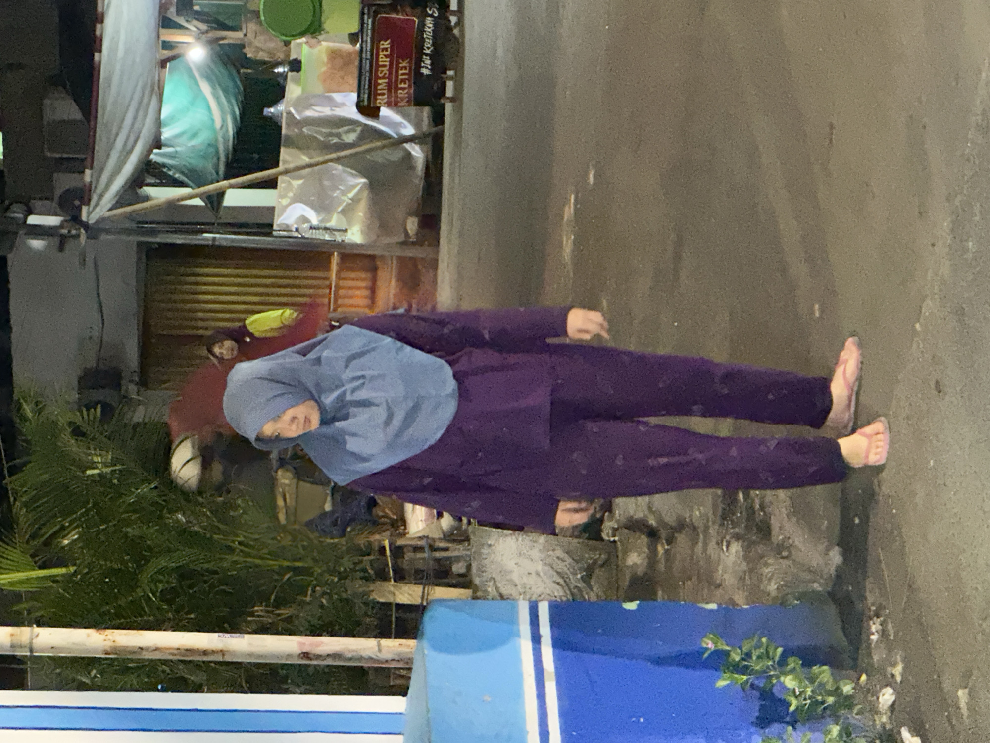 The image shows a woman standing on a wet street in North Jakarta, Indonesia. She is wearing a purple long-sleeved shirt and pants with a light blue hijab. Her bare feet are visible. The background includes a partially open metal rolling door, a building with a tarp overhead, and a sign that reads "RUM SUPER KRETEK." There is a blue and white structure in the foreground, possibly a part of a building or street furniture. The overall setting is urban and the wet street suggests it has recently rained.