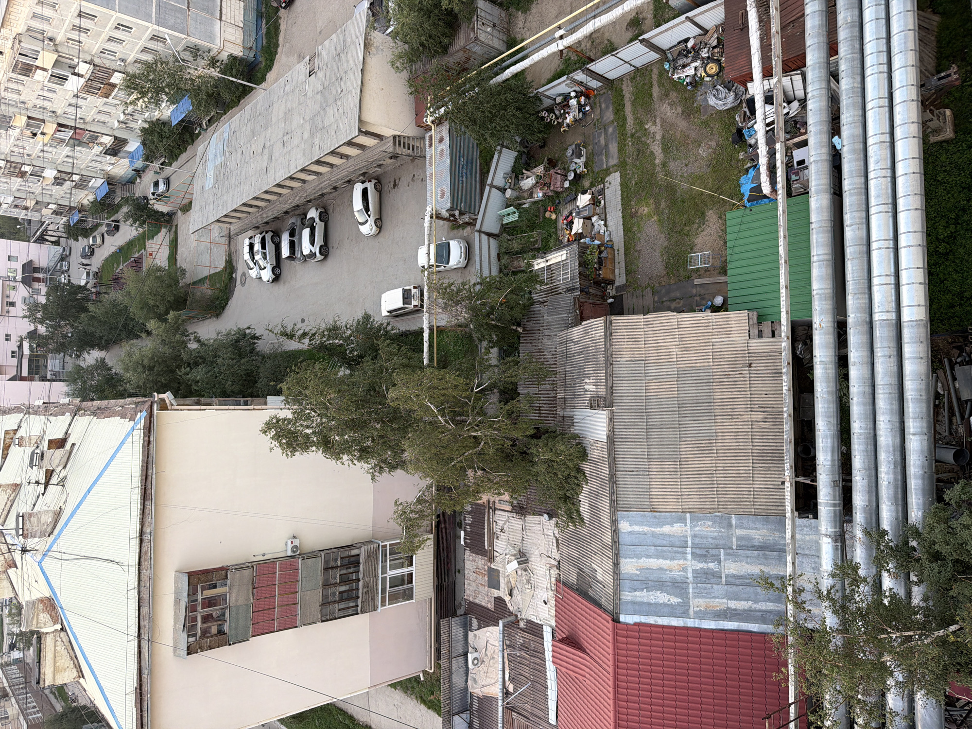 This is an aerial, high-angle, daytime, medium shot of a residential area in Yakutsk, Russia, likely taken from a window of a multi-story building. In the foreground, several large, parallel, metallic pipes run horizontally across the frame. Behind these pipes, there are several single-story structures with corrugated metal roofs, some red, some silver and rusted, and one green. One of these structures appears to be a small garage or workshop. Adjacent to these structures is a dirt yard with scattered debris including piles of junk, scrap metal, and what appears to be an old yellow tractor.
Further back and to the left, a large, light-colored, multi-story apartment building is visible, with a light-colored corrugated metal roof and a protruding, multi-paneled balcony. To the right of this apartment building, another, much larger, grey, multi-story apartment building dominates the middle ground, with many windows and balconies. A paved walkway runs between these buildings, with several parked cars visible. Trees and greenery are interspersed throughout the scene, with a large birch tree leaning significantly over some of the single-story structures. The environment appears somewhat utilitarian and weathered.