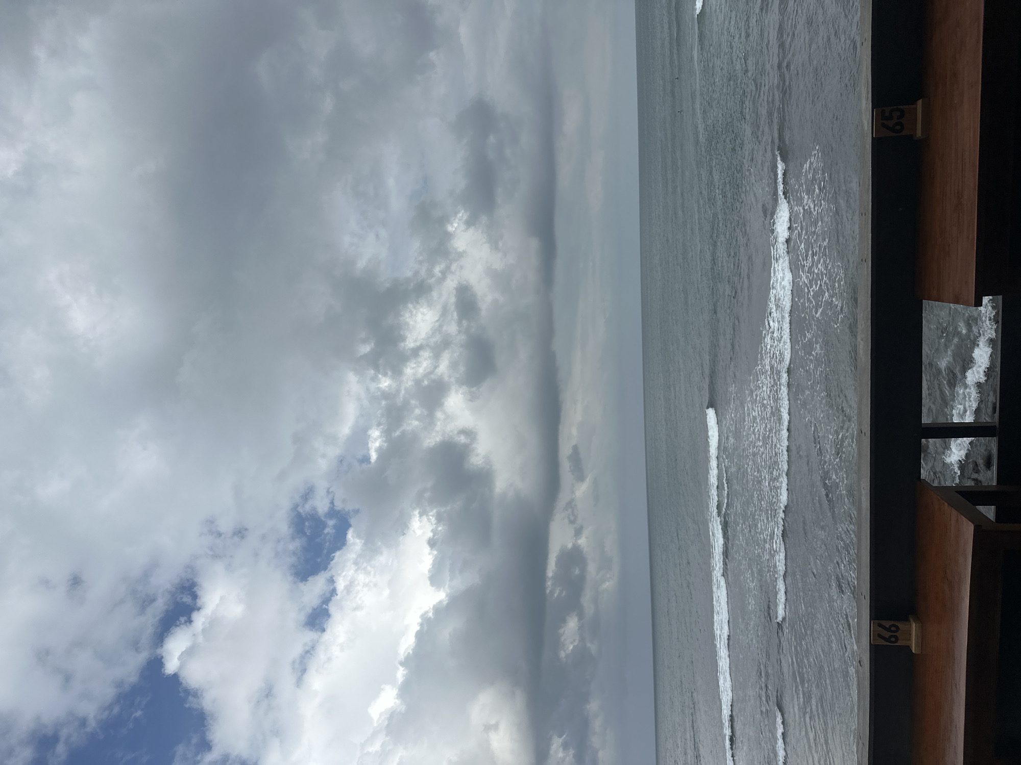 The image depicts a serene beach scene in North Kuta, Indonesia. The sky is filled with dramatic, heavy clouds, suggesting an overcast or possibly stormy day. The sea is calm with gentle waves rolling towards the shore. The foreground features wooden structures, likely part of a beachside cafe or viewing platform, with visible numbering: "65" and "66" are clearly discernible on wooden blocks. These numbers might indicate beach access points or seating arrangements. The overall mood is peaceful and somewhat melancholic due to the cloudy weather. The time of day is difficult to pinpoint precisely, but the diffused light suggests it could be morning or late afternoon. There are no people visible in the frame, emphasizing the solitude of the scene. The focus is on the natural elements of the ocean and sky, contrasted with the man-made structures that blend into the environment.